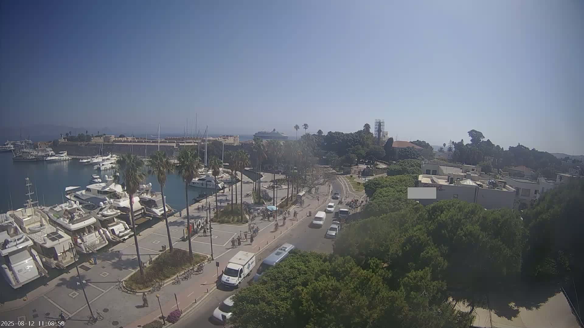 An aerial view shows a harbor filled with yachts and a cruise ship in the distance on a clear, sunny day, with palm trees and pedestrians along the waterfront.