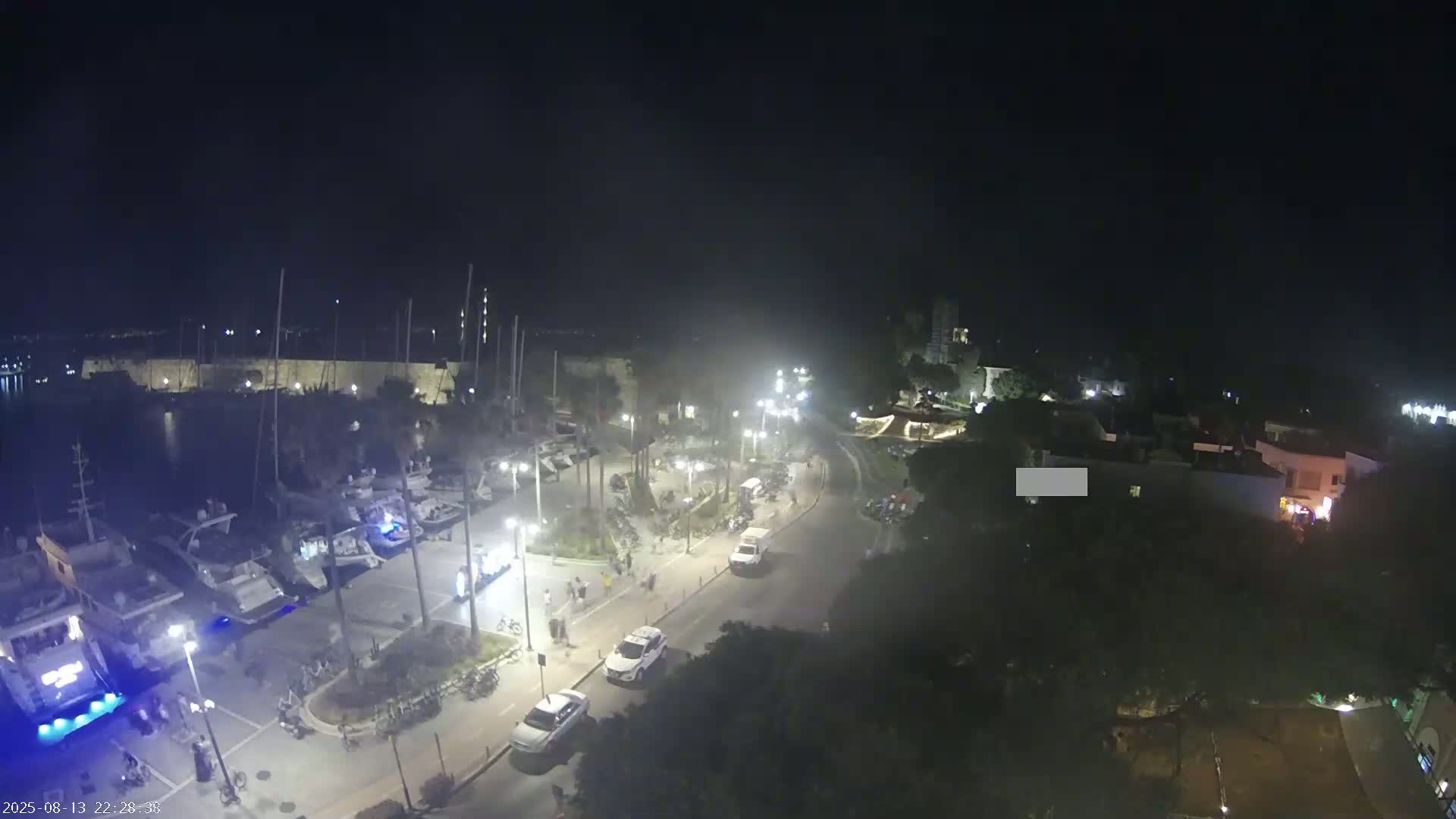 An elevated nighttime view shows a harbor with moored boats, a town square with people and vehicles, and surrounding buildings under a clear, dark sky.