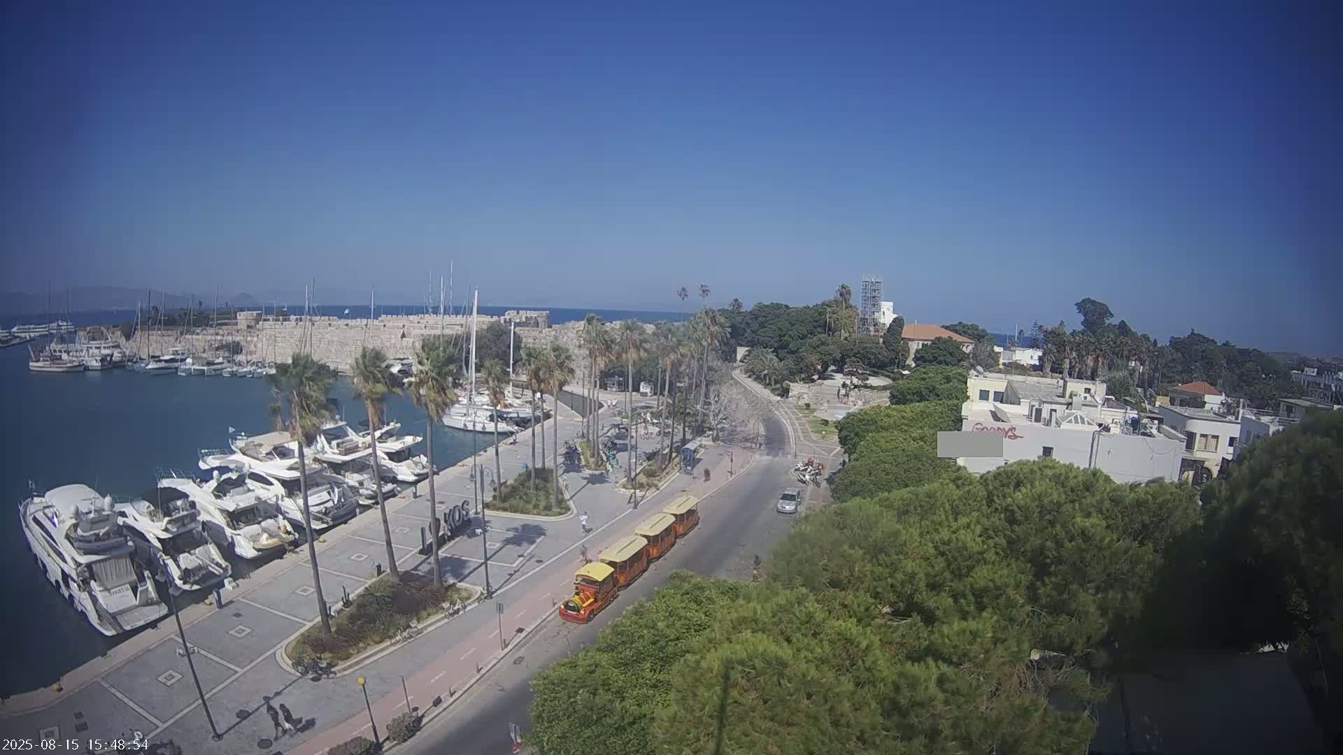 A harbor filled with boats is seen from an elevated viewpoint on a sunny day, with palm trees lining a paved area next to a road where a tourist train is traveling.