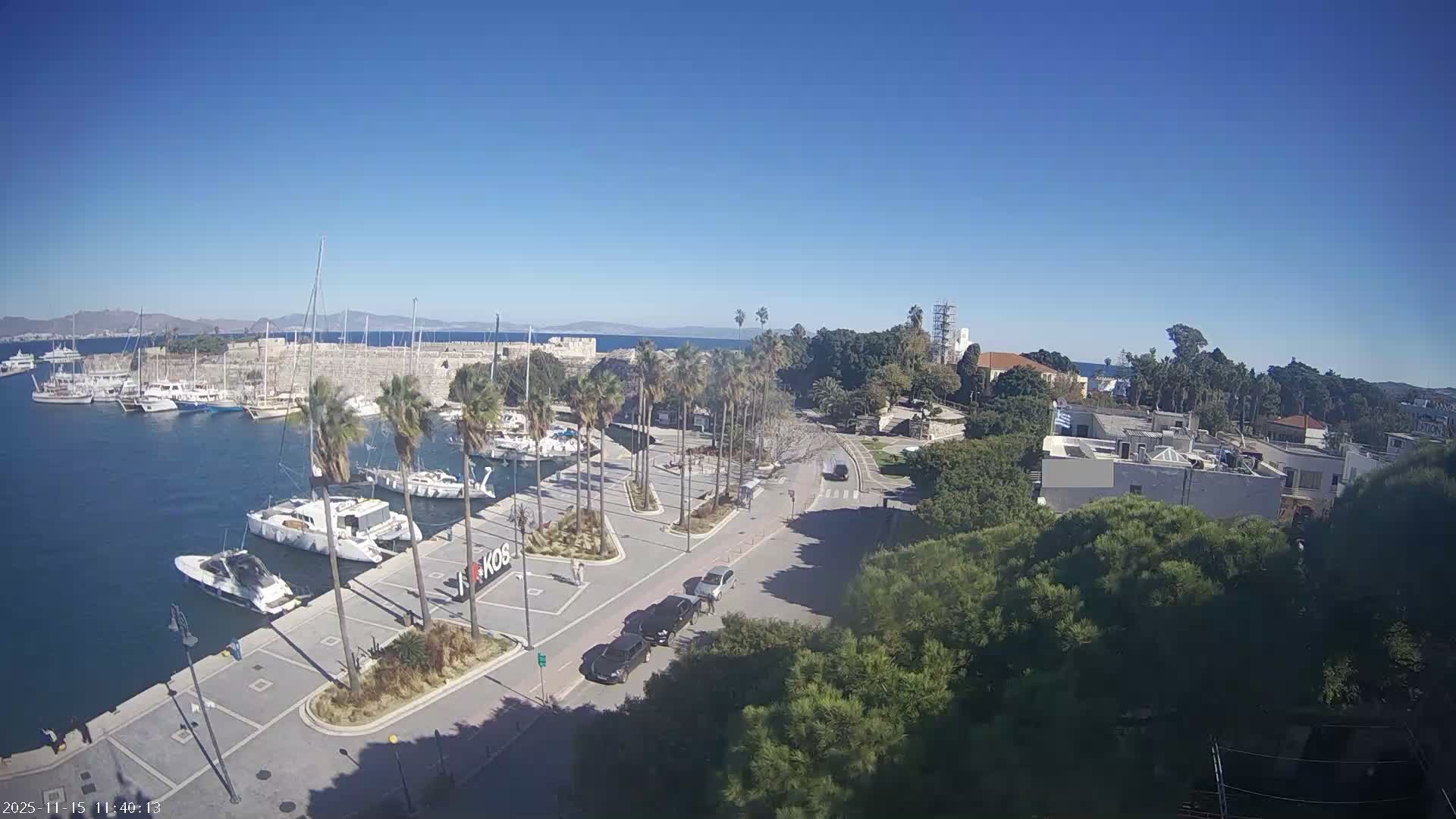 On a clear and sunny day, an elevated view captures a lively harbor filled with docked boats and yachts, a palm-tree-lined promenade with a road and cars, historic stone fortifications, and a vibrant coastal town stretching towards distant mountains under a bright blue sky.