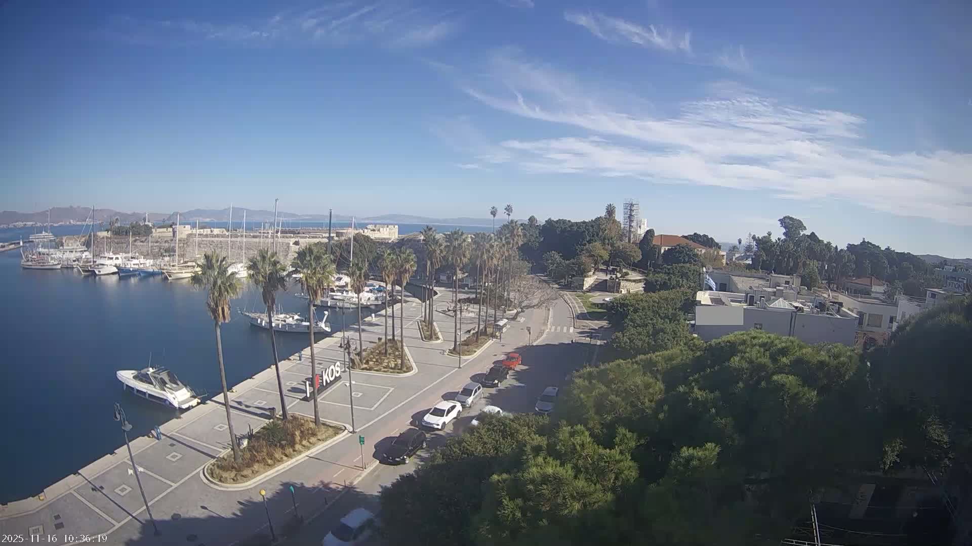 A sunny day with a clear blue sky and scattered wispy clouds overlooks a harbor filled with numerous boats, a street lined with palm trees and several cars, and a coastal town featuring buildings, lush greenery, and distant mountains.