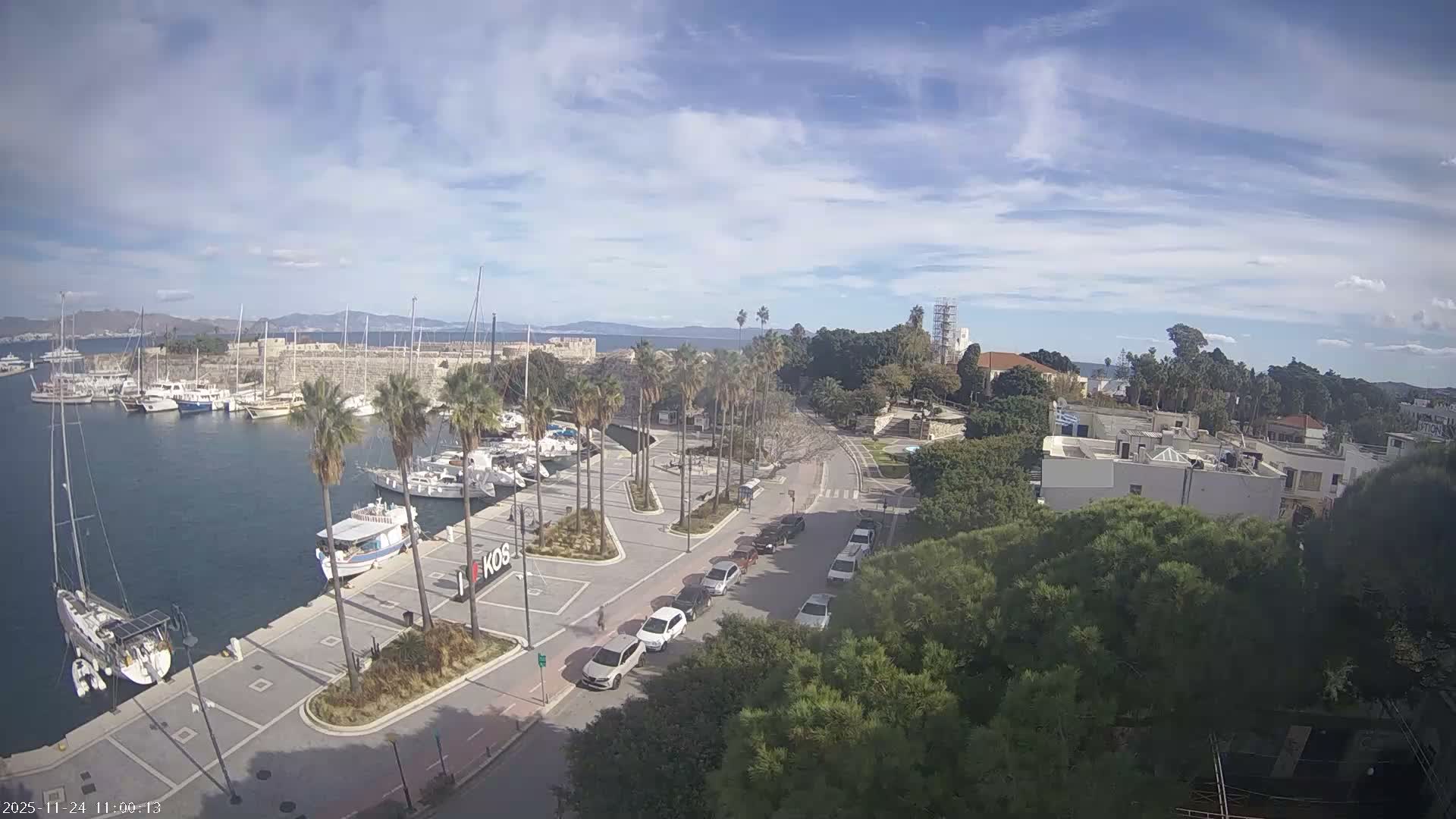 An elevated view reveals a bustling harbor filled with boats alongside a tree-lined street with parked cars, surrounded by buildings and distant mountains, all under a partly cloudy sky.