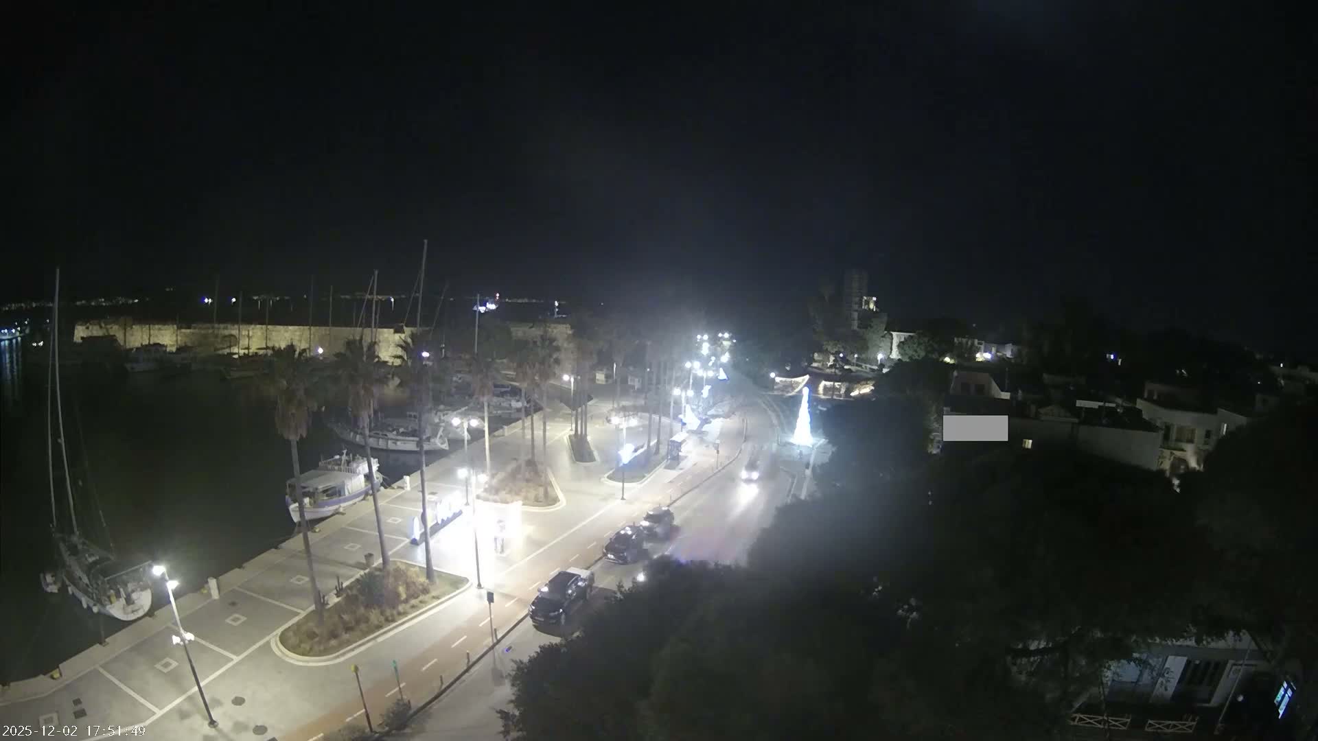 A clear night scene showcases a bustling harbor with boats, a brightly lit promenade lined with palm trees and streetlights, moving vehicles on a road, and an illuminated Christmas tree, all set against distant buildings under a dark sky.