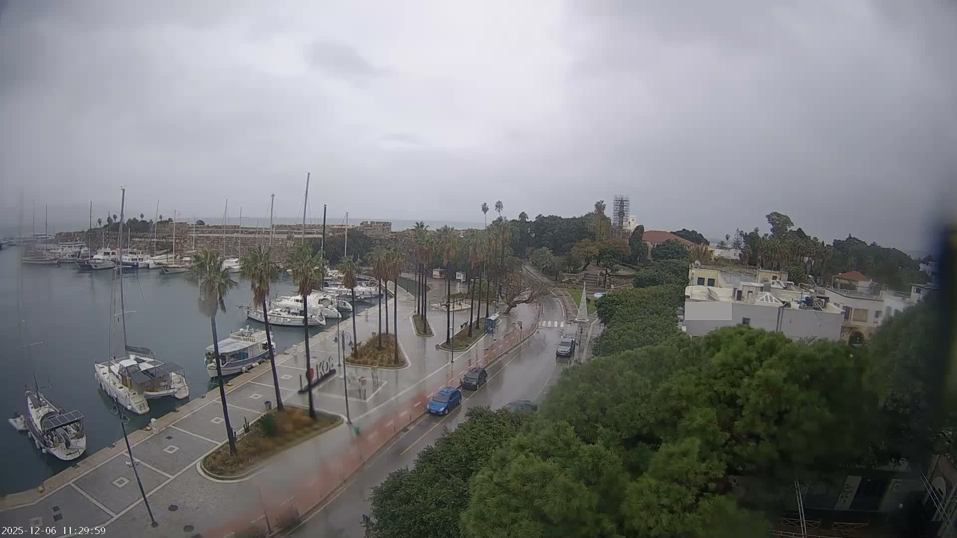 An aerial view captures a marina full of boats, a wet road with scattered cars, and palm trees lining the waterfront promenade, all under an overcast and rainy sky with historical fortifications and buildings in the distance.