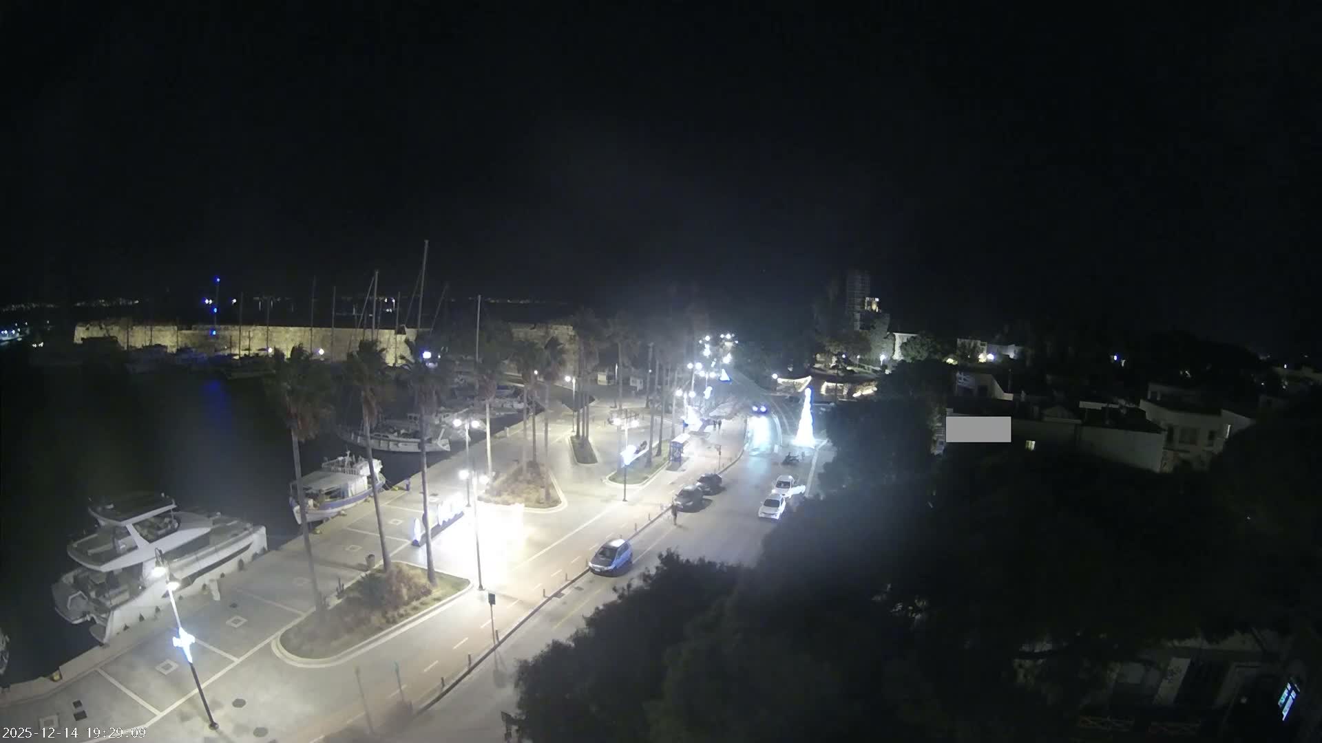 An aerial view captures a marina full of boats, a wet road with scattered cars, and palm trees lining the waterfront promenade, all under an overcast and rainy sky with historical fortifications and buildings in the distance.