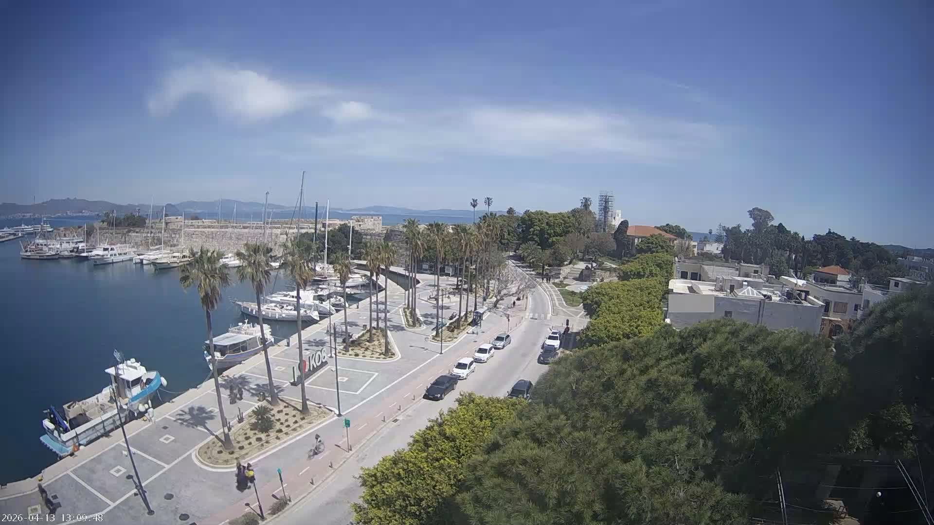 An elevated view shows a harbor filled with boats and yachts, a palm-lined promenade with people and cars, and a city extending into the distance under a partly cloudy sky.