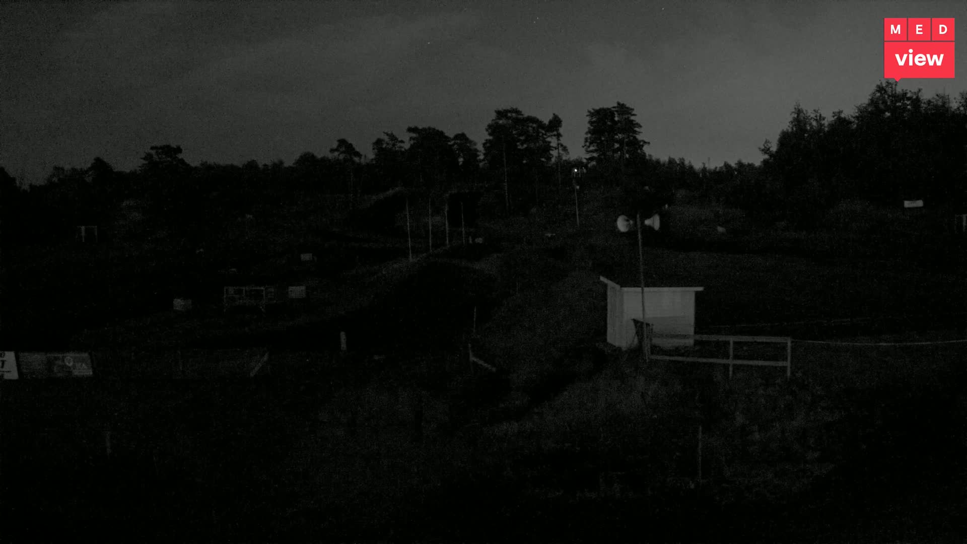 A nighttime, low-light view of a dark, open area with a few small structures and a line of trees in the background under a mostly clear sky.