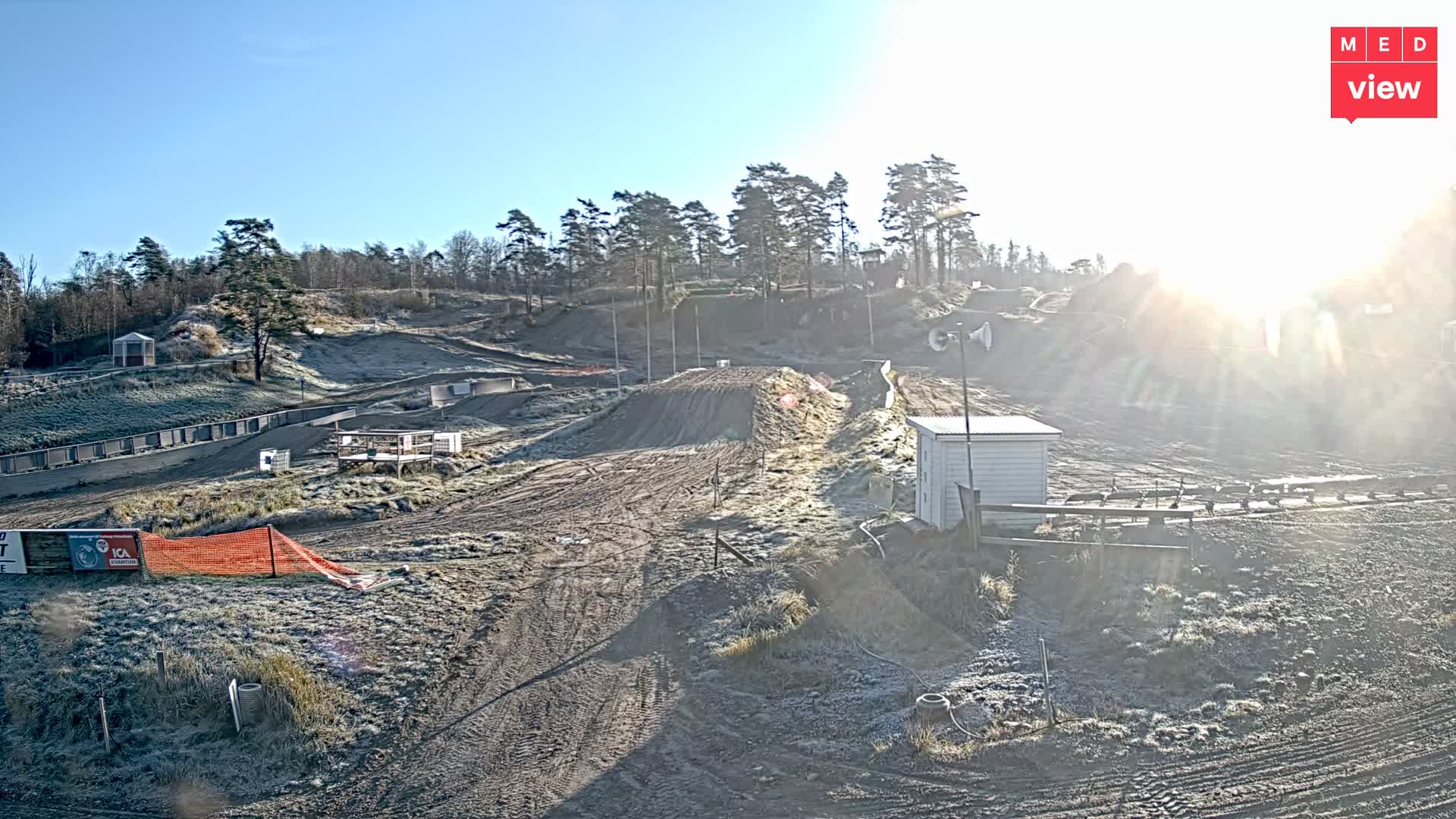 A frosted dirt track with several ramps and hills, possibly for motocross, lies under a clear, sunny sky with bright low-angle sunlight creating a glare on the right.