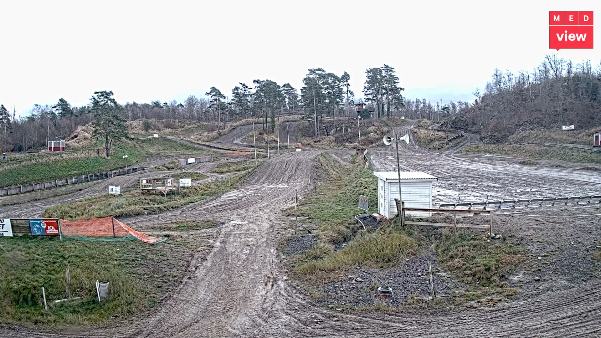 A muddy, winding outdoor motocross track with jumps, scattered small buildings, and spectator areas stretches across a hilly landscape with mostly bare trees under a dull, overcast sky.