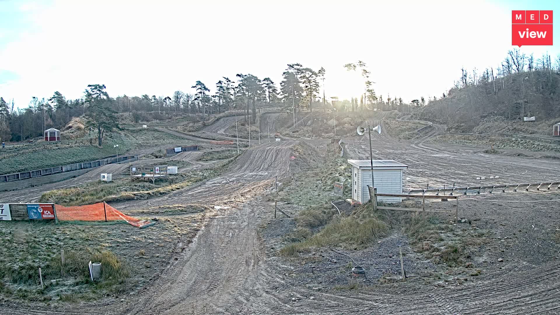 A muddy, winding outdoor motocross track with jumps, scattered small buildings, and spectator areas stretches across a hilly landscape with mostly bare trees under a dull, overcast sky.