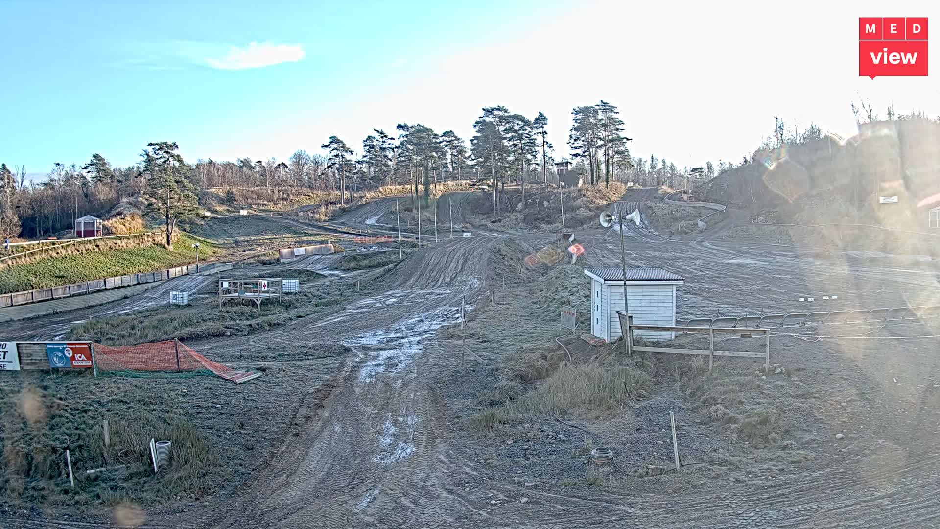 A muddy, winding outdoor motocross track with jumps, scattered small buildings, and spectator areas stretches across a hilly landscape with mostly bare trees under a dull, overcast sky.