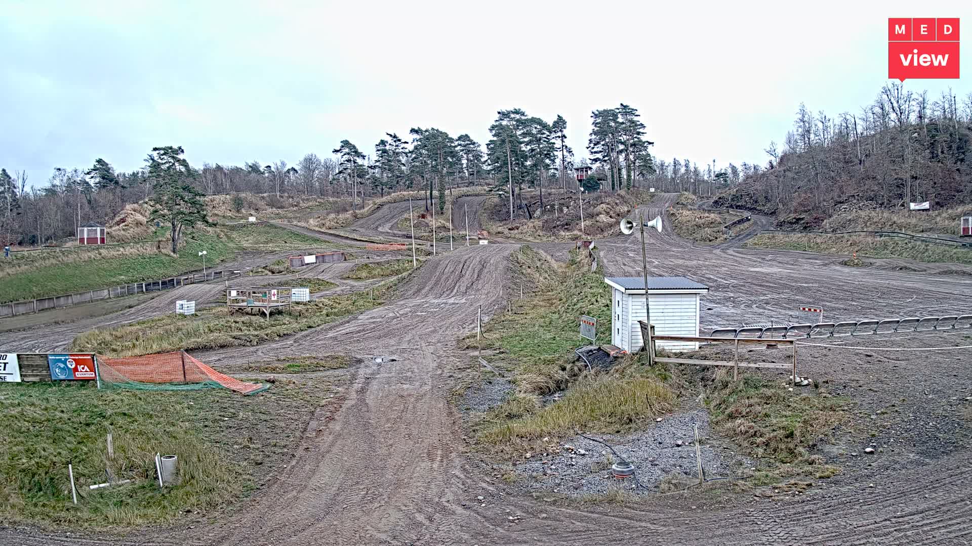 A wide view captures a muddy dirt track with numerous turns and elevation changes, indicative of a motocross or off-road course, surrounded by sparse, wintery trees and grassy slopes under an overcast sky.