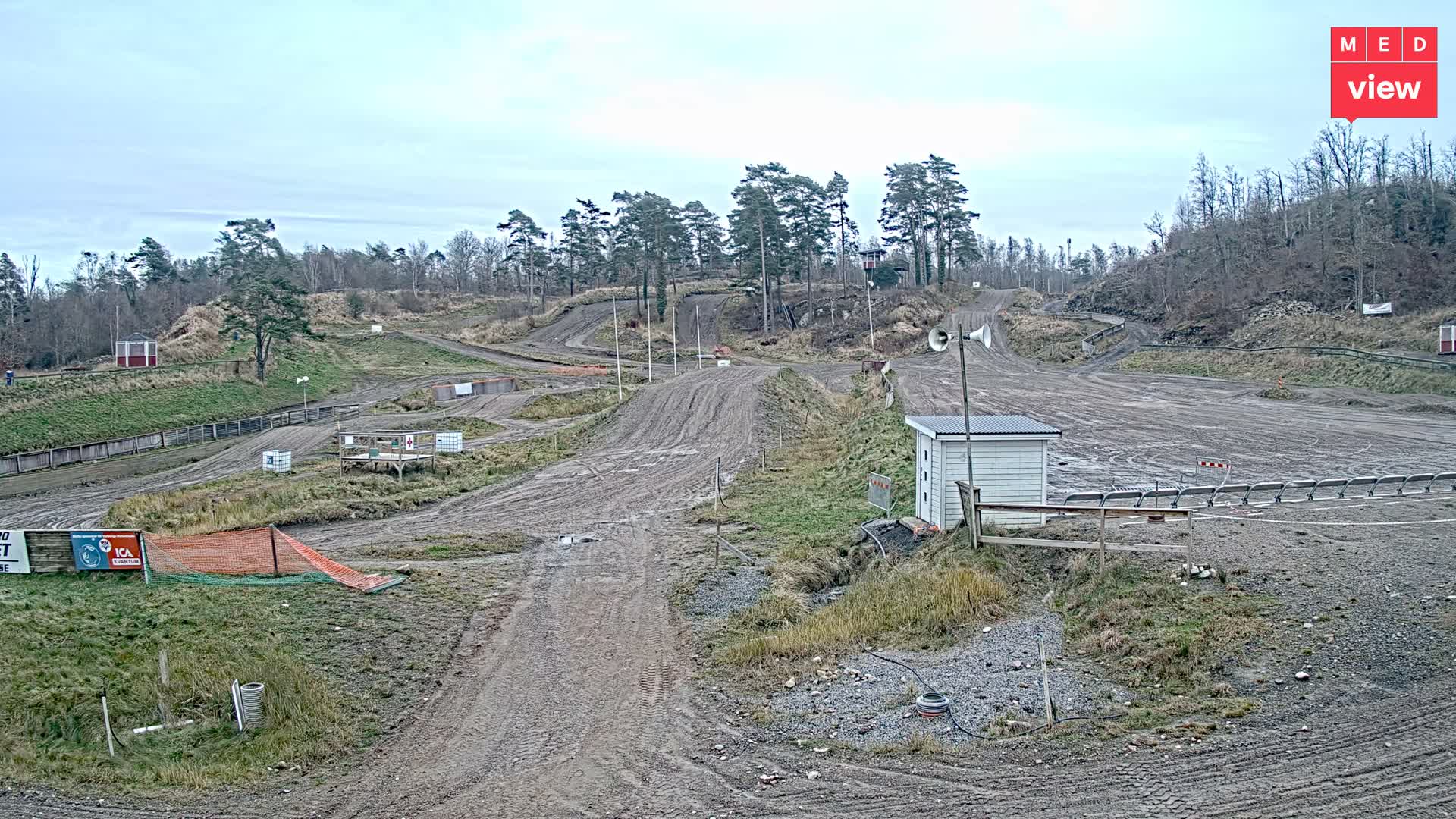 A muddy motocross track with winding dirt paths and jumps is seen on an overcast and damp day, surrounded by hills covered in a mix of bare deciduous and evergreen trees, with a small white building and loudspeakers in the foreground.