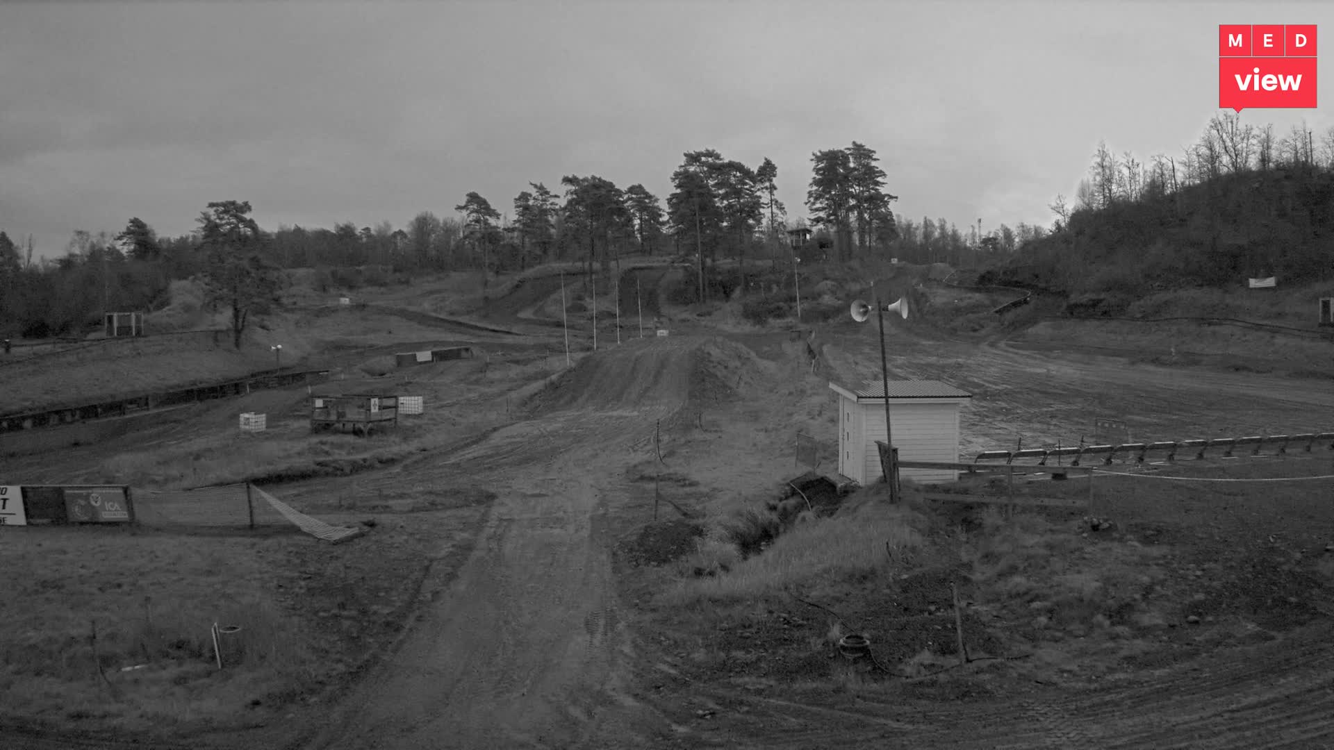 A rugged motocross track featuring dirt hills, winding paths, scattered trees, and a small white building with loudspeakers is visible under an overcast and gloomy sky.