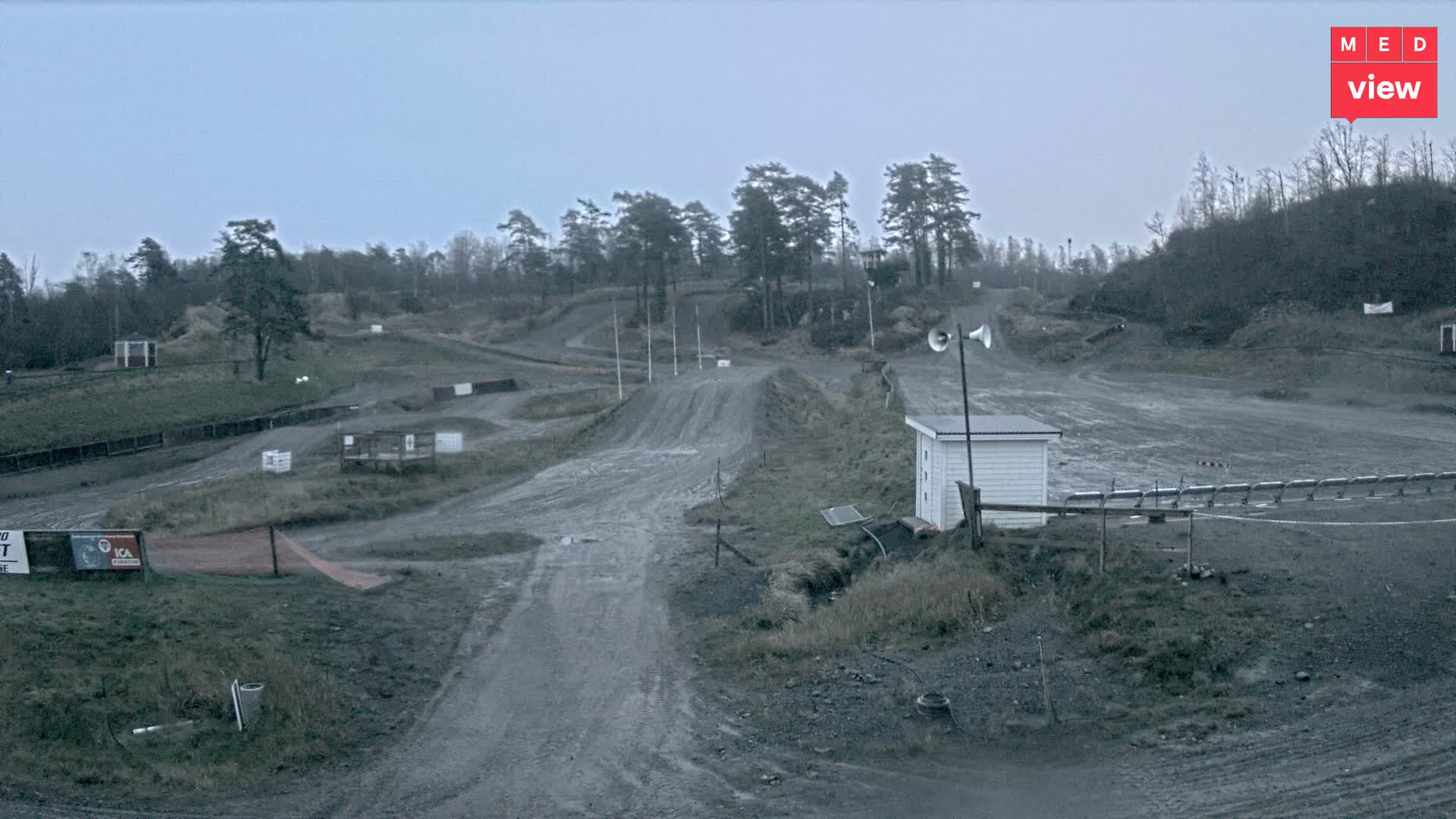 A rugged motocross track featuring dirt hills, winding paths, scattered trees, and a small white building with loudspeakers is visible under an overcast and gloomy sky.