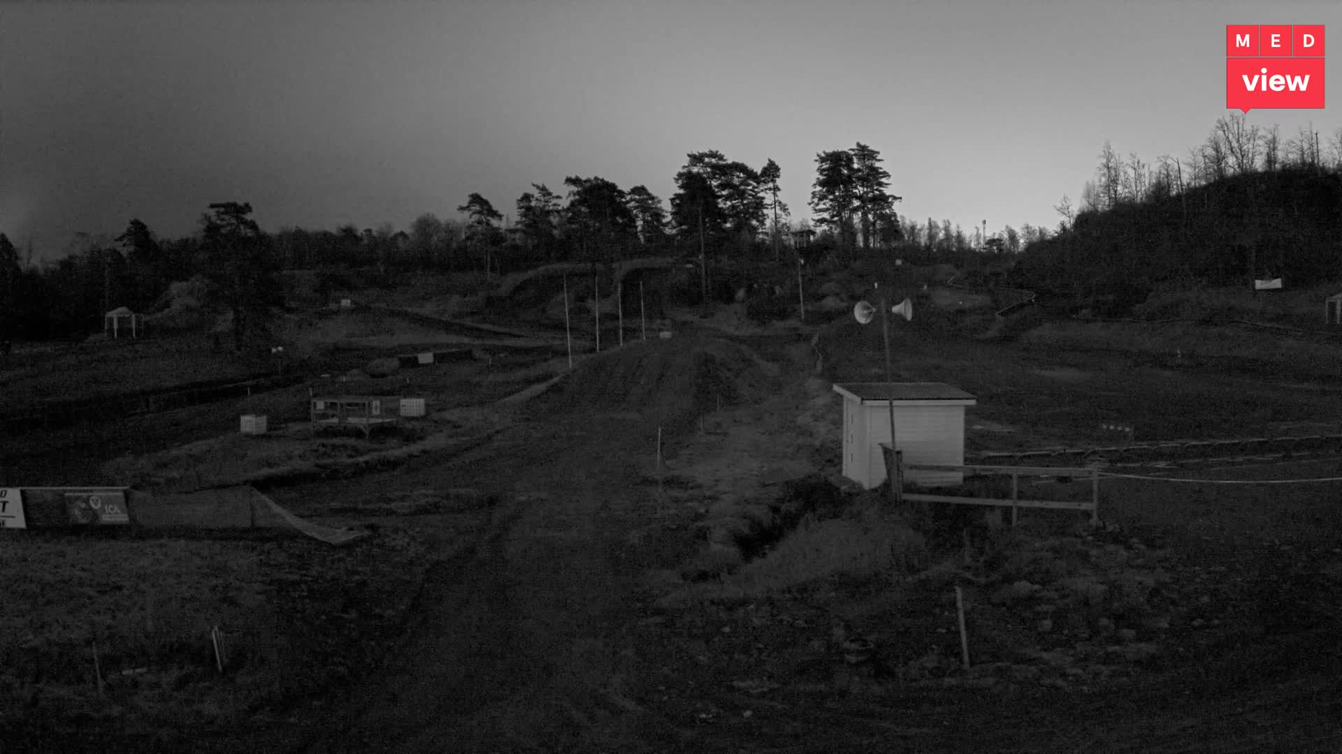 A dimly lit, hilly outdoor training range featuring dirt paths, sparse trees, a small white building with large speakers, and various targets under a twilight sky.