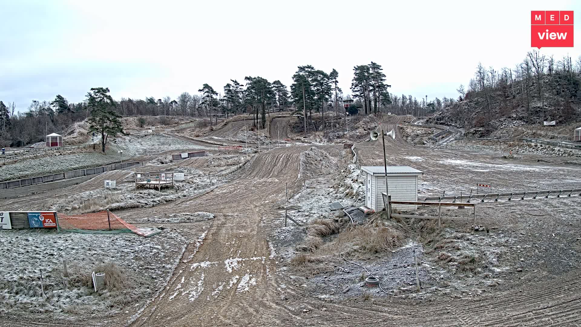 A dimly lit, hilly outdoor training range featuring dirt paths, sparse trees, a small white building with large speakers, and various targets under a twilight sky.