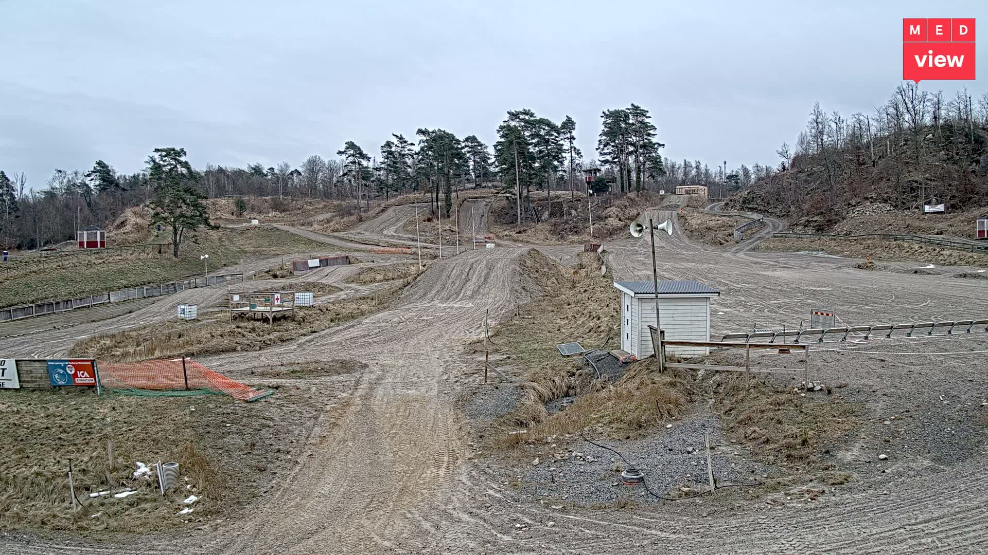 An empty motocross track with numerous dirt ramps and turns, surrounded by bare trees and dry grass, is pictured under a dull, overcast sky, featuring a small white building with loudspeakers and hints of lingering snow.
