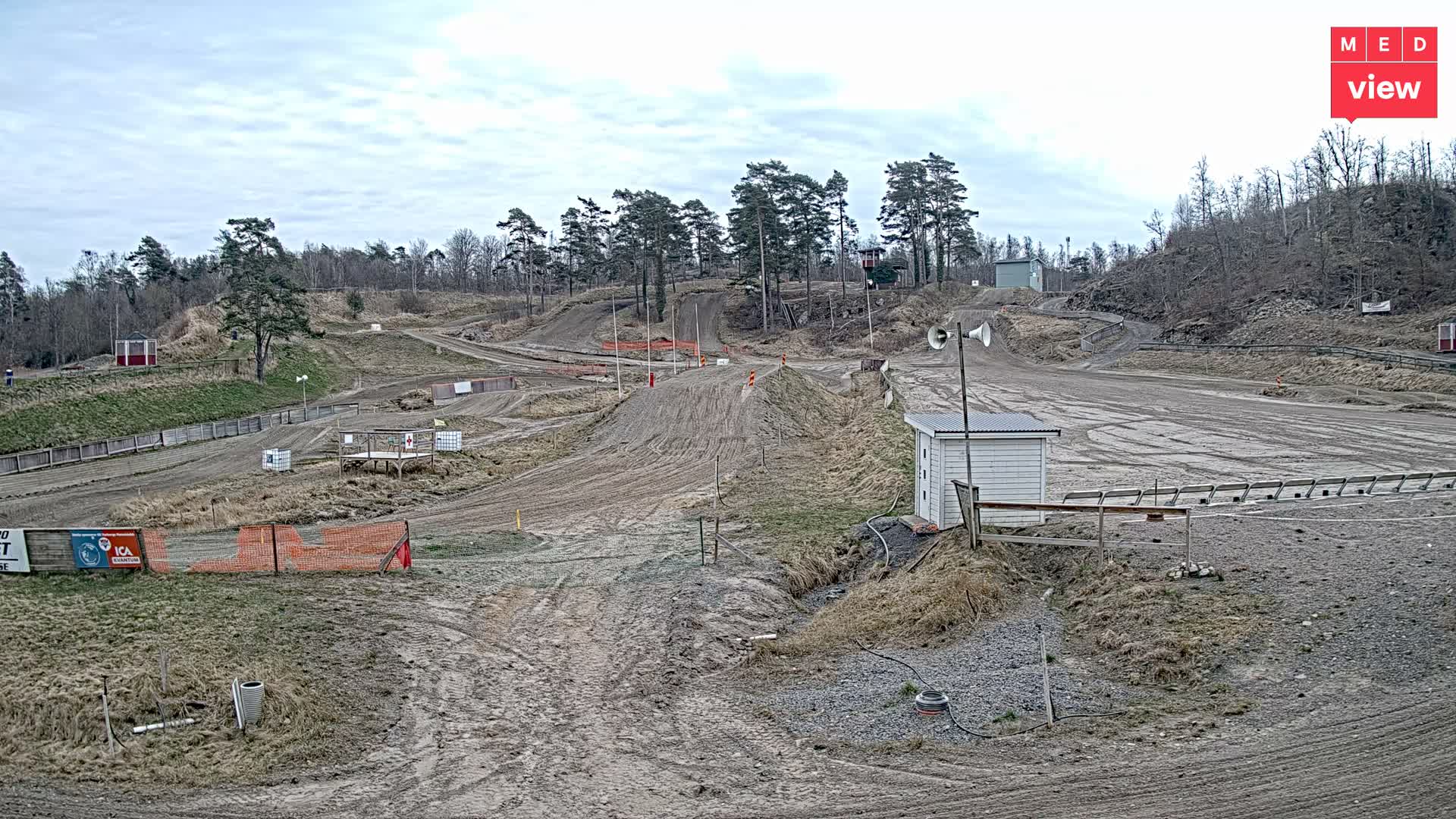An empty motocross track with numerous dirt ramps and turns, surrounded by bare trees and dry grass, is pictured under a dull, overcast sky, featuring a small white building with loudspeakers and hints of lingering snow.