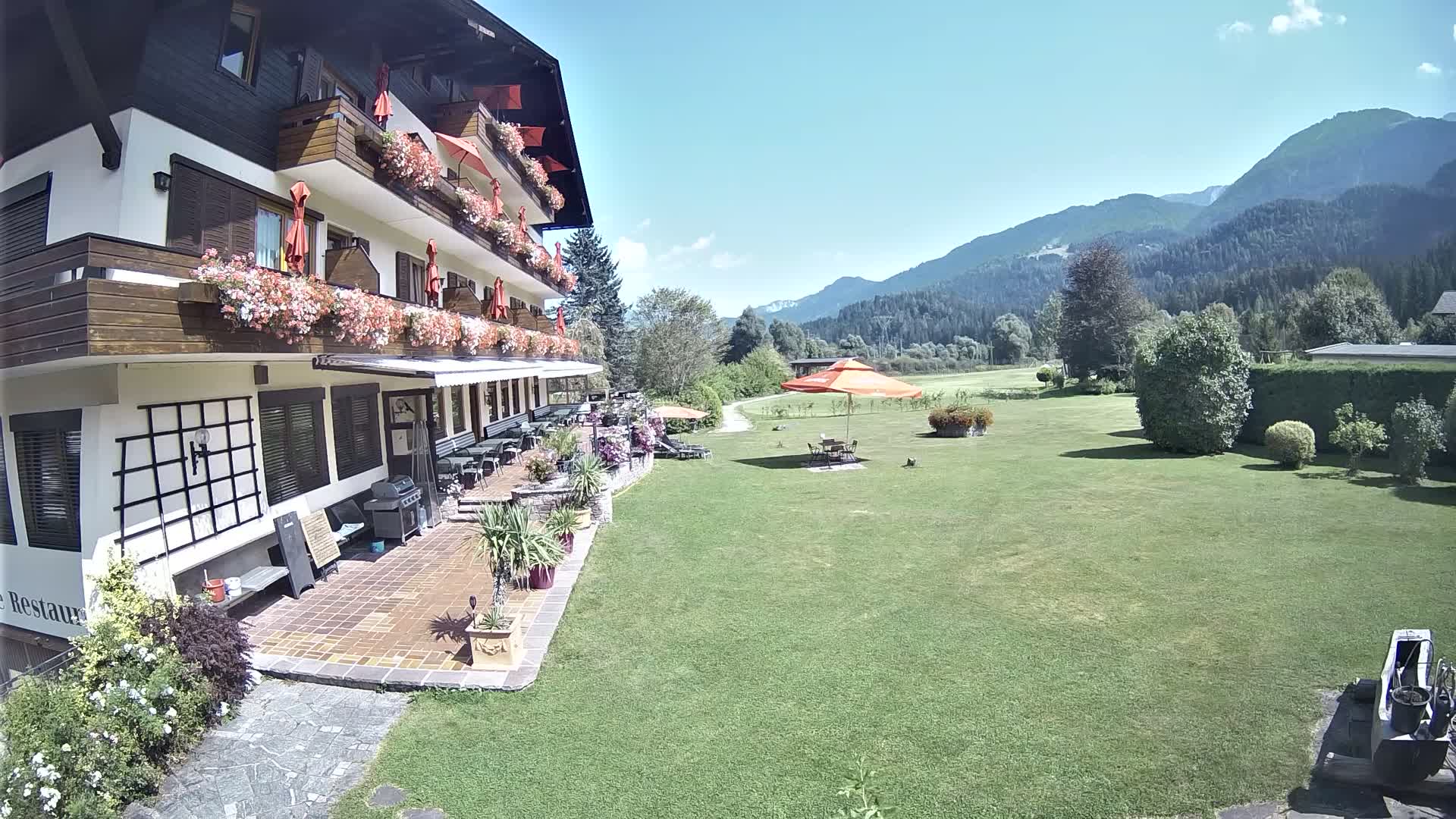 A multi-story building with balconies and patio furniture overlooks a large, grassy lawn with an umbrella-shaded table and chairs, all under a sunny, hazy sky with mountains in the background.