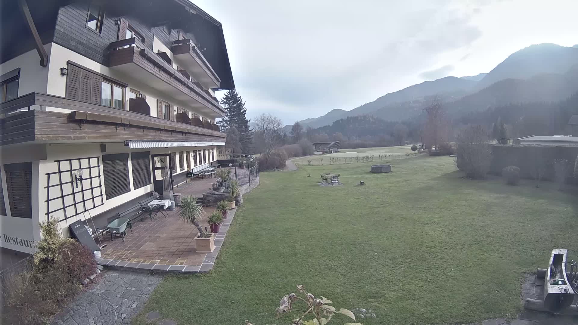 A multi-story traditional building with wooden balconies and a tiled patio offering outdoor seating faces an expansive green lawn, featuring a well and distant golf course elements, all set against a backdrop of mountains under a grey, overcast sky.