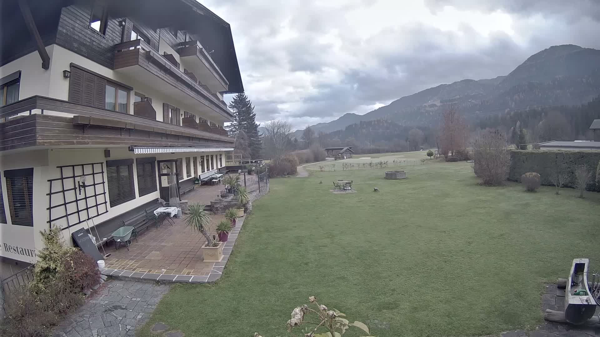 A multi-story building with a patio and balconies overlooks a wide green lawn backed by tree-covered mountains, all under a heavily overcast sky.