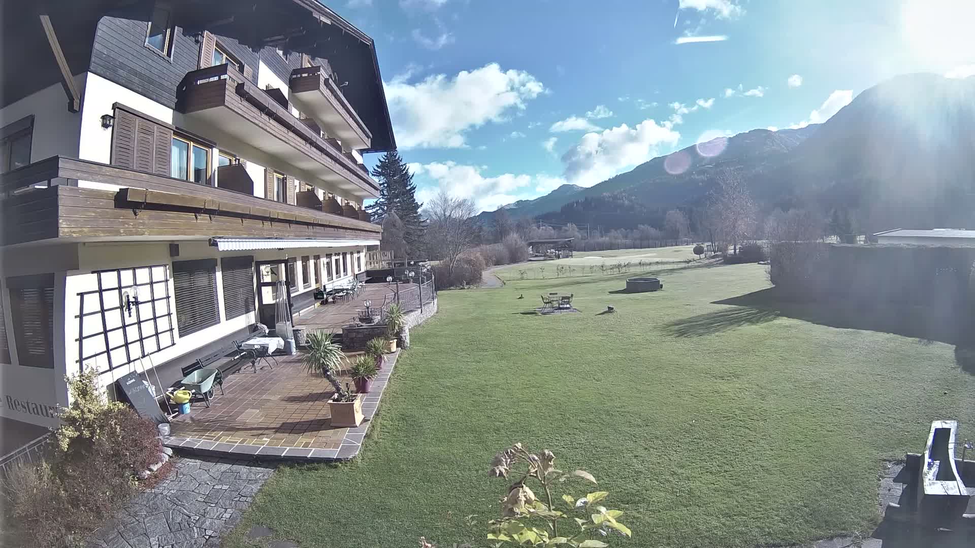A multi-story building with a patio and balconies overlooks a wide green lawn backed by tree-covered mountains, all under a heavily overcast sky.