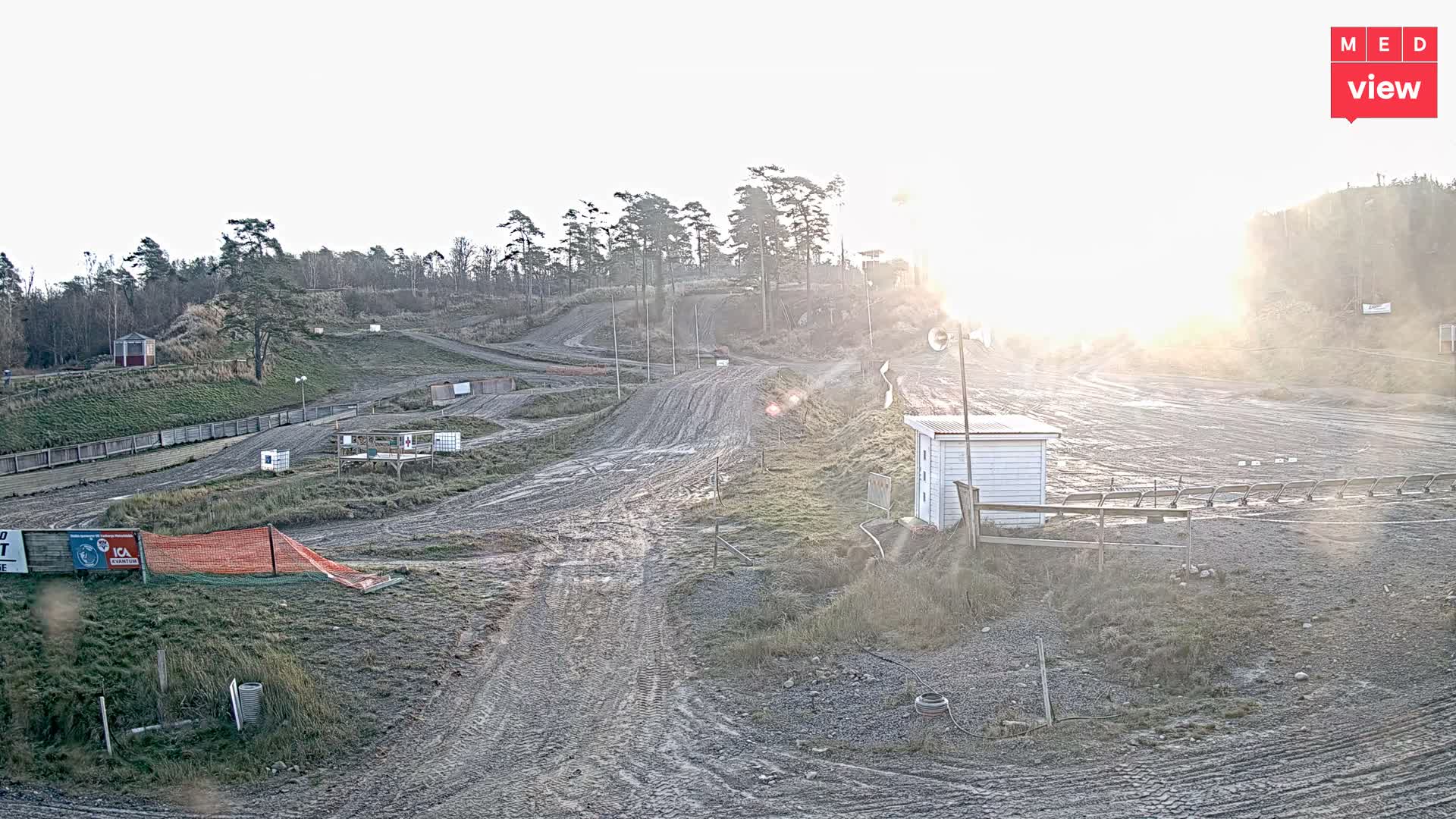 A winding dirt track course with multiple paths and patches of sparse grass and trees spans rolling hills, dotted with small buildings, under a bright, low sun causing significant glare.