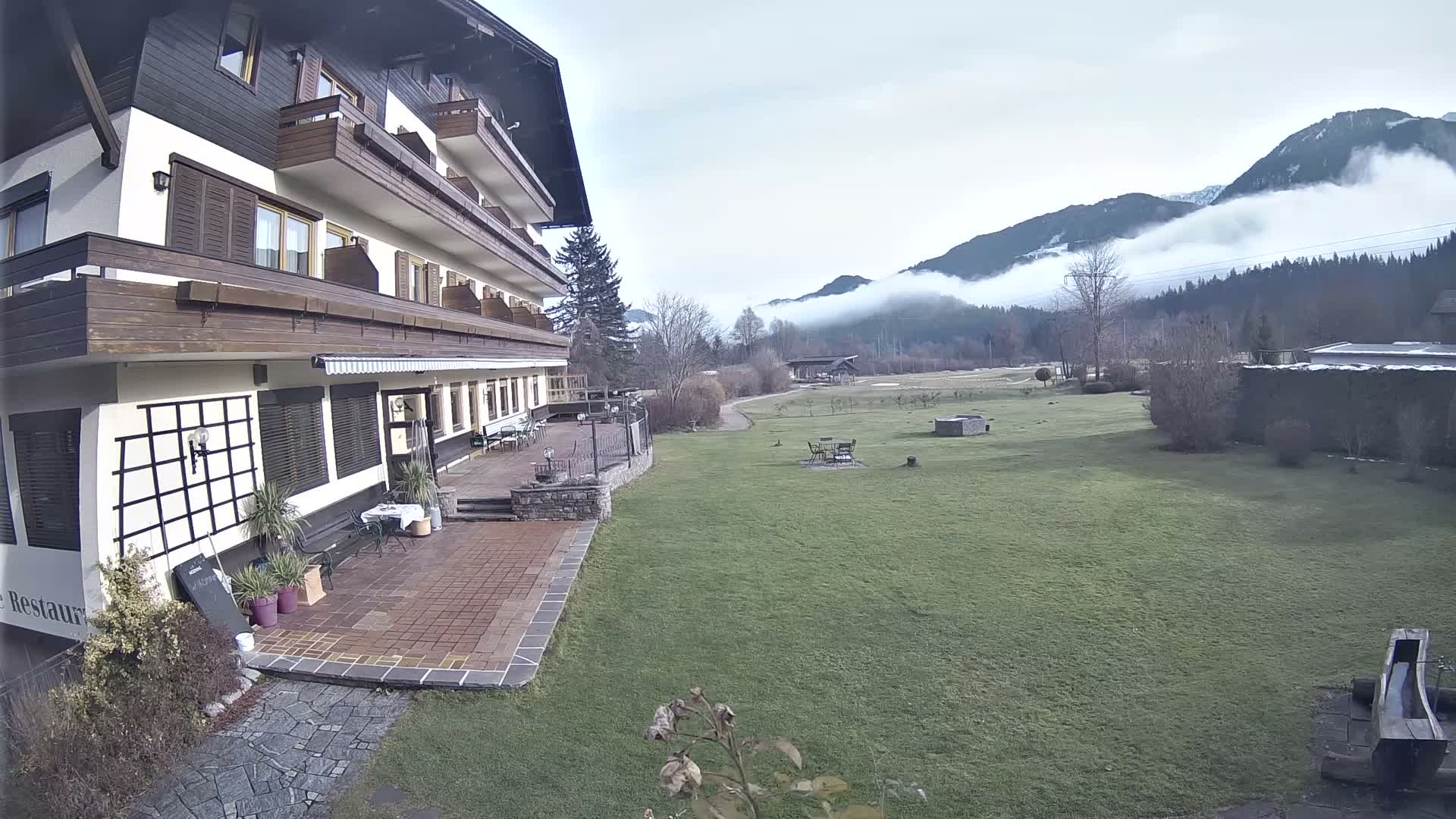 A winding dirt track course with multiple paths and patches of sparse grass and trees spans rolling hills, dotted with small buildings, under a bright, low sun causing significant glare.