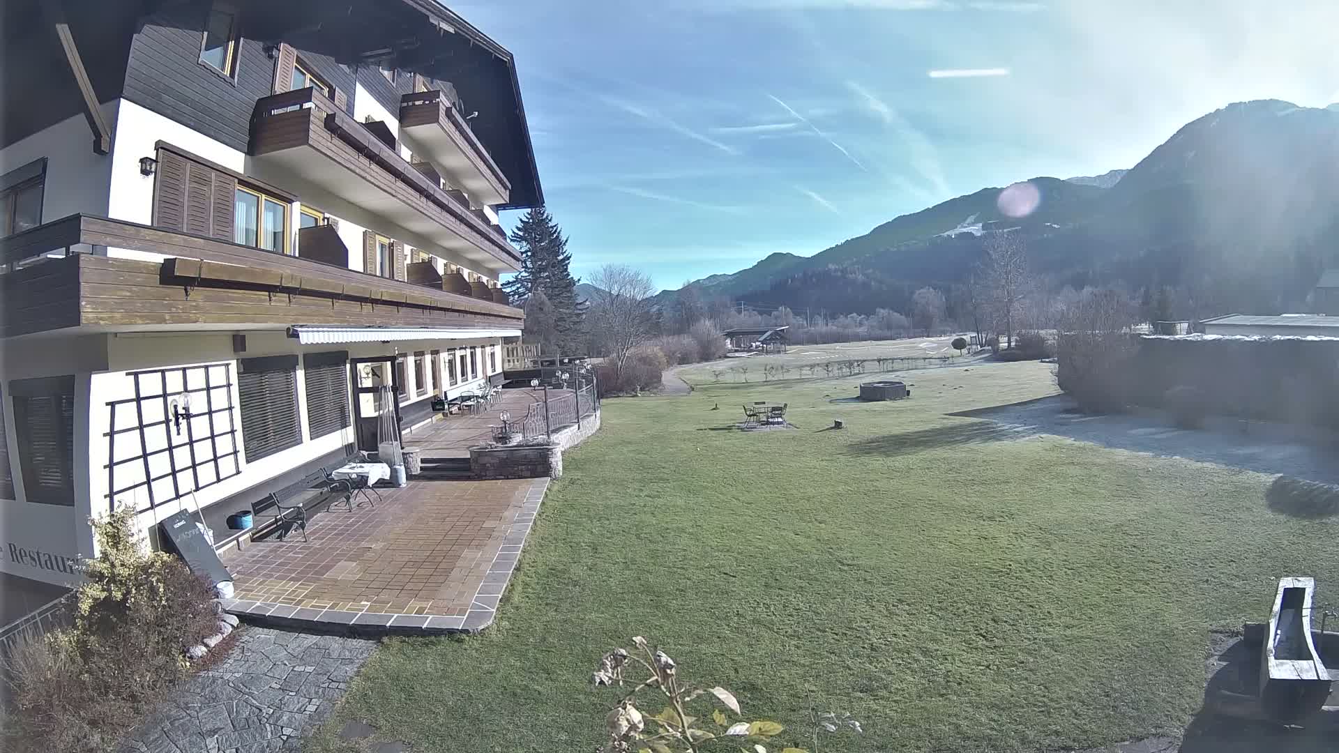 A multi-story building with balconies and an outdoor patio fronts a wide green lawn with scattered frost, leading to a valley and mountains under a clear, sunny sky.