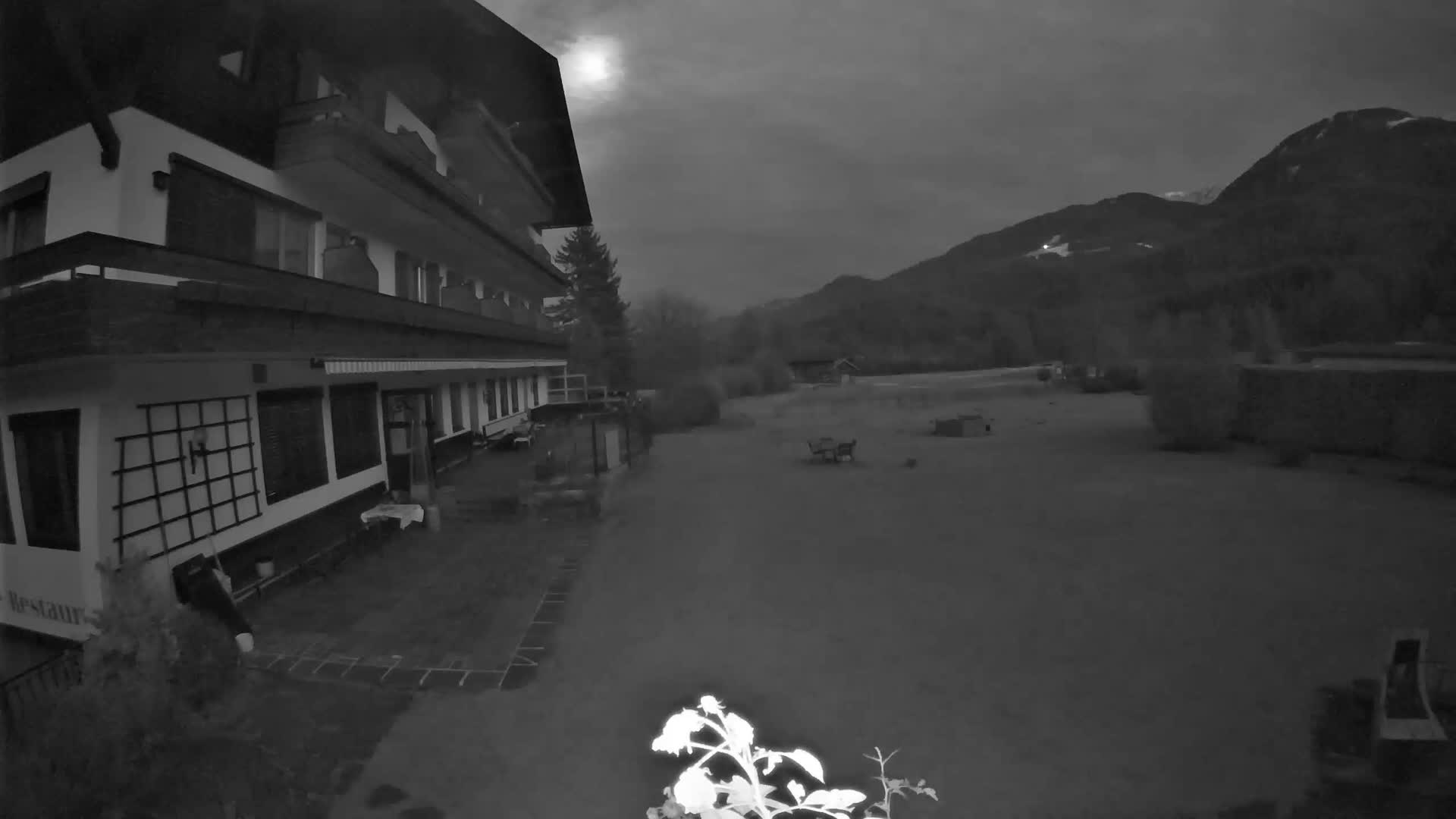 An outdoor scene reveals a multi-story building beside a grassy field with scattered furniture and a brightly glowing plant in the foreground, all set against a backdrop of mountains under an overcast night sky with a hazy moon.