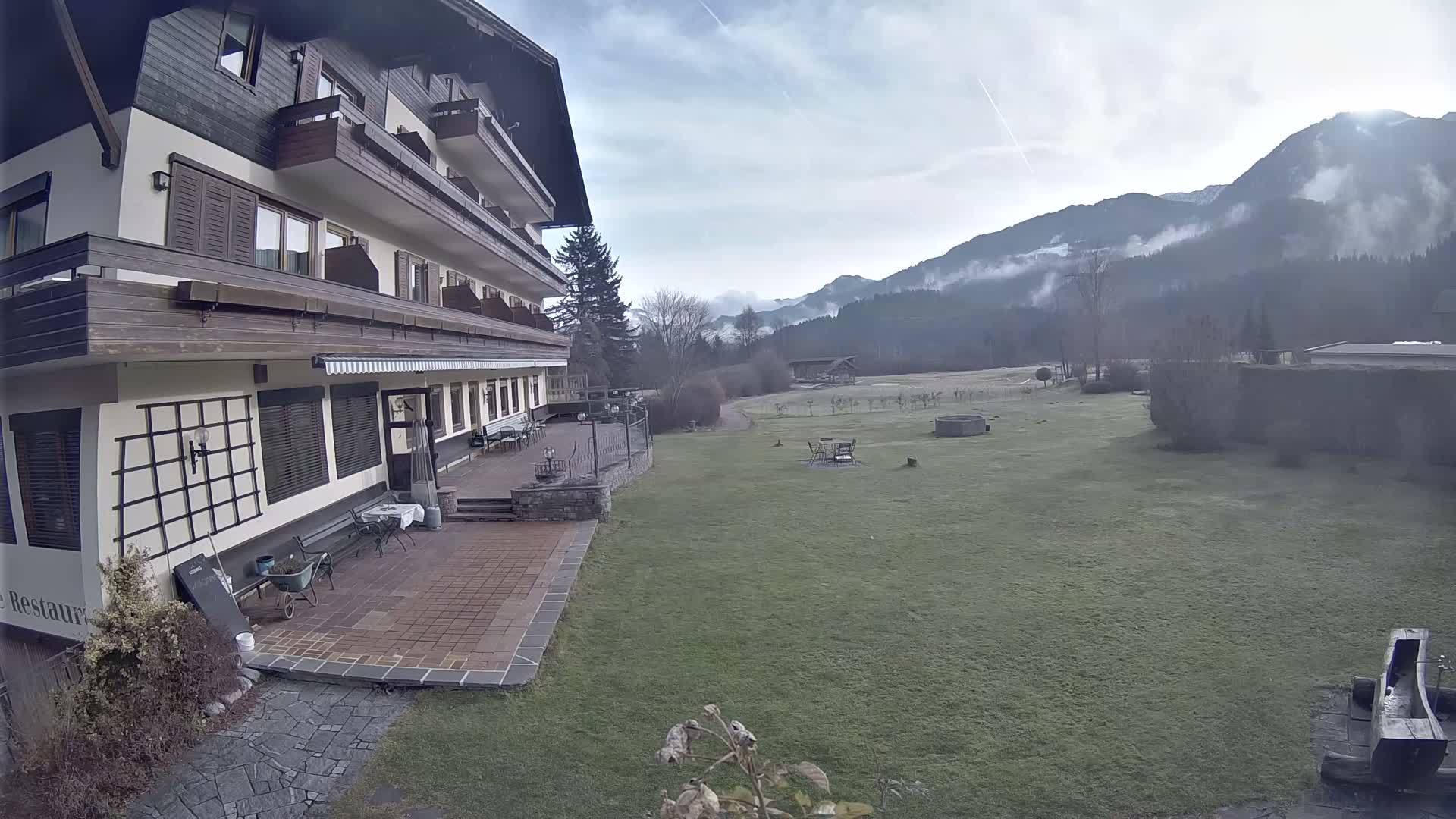 A multi-story building with balconies and a paved patio overlooks a vast green lawn, beyond which misty, tree-covered mountains rise under a bright, cloudy sky.
