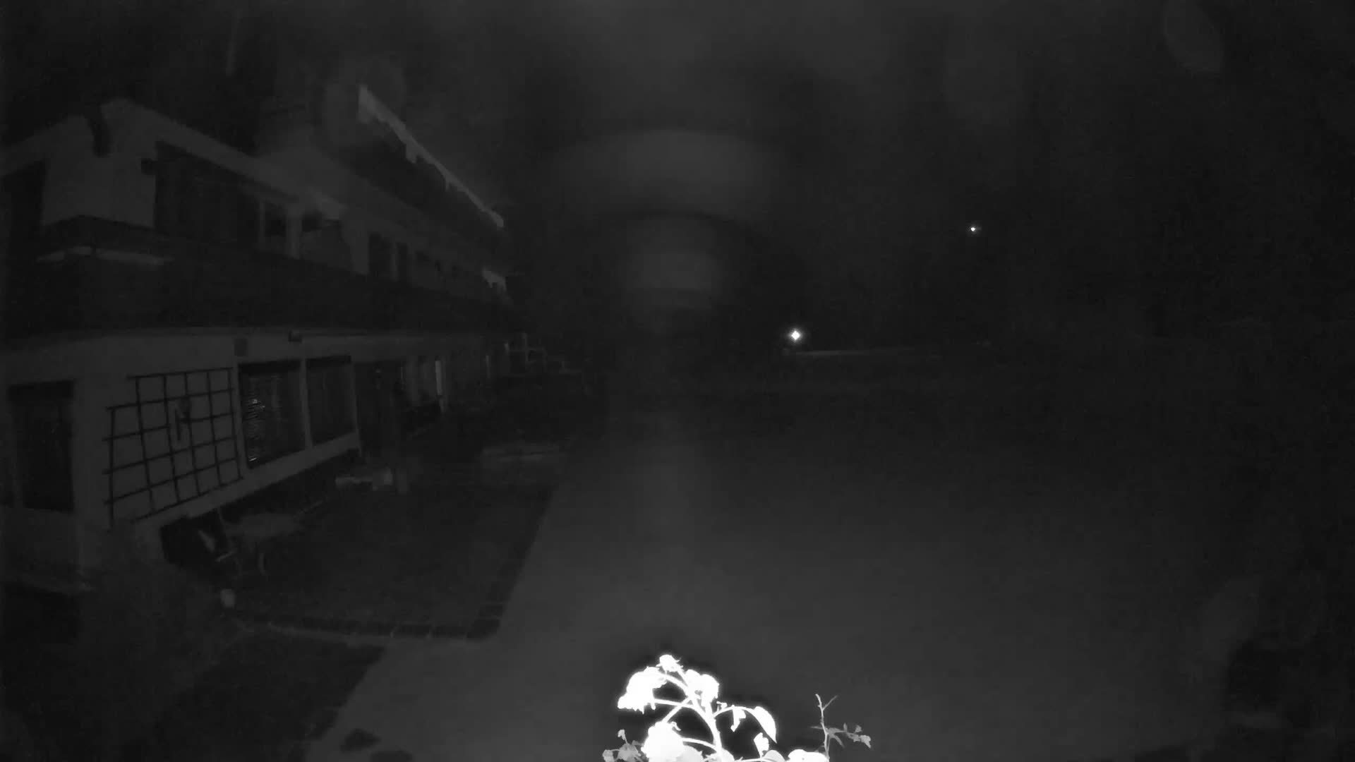 A multi-story building with balconies and a paved patio overlooks a vast green lawn, beyond which misty, tree-covered mountains rise under a bright, cloudy sky.