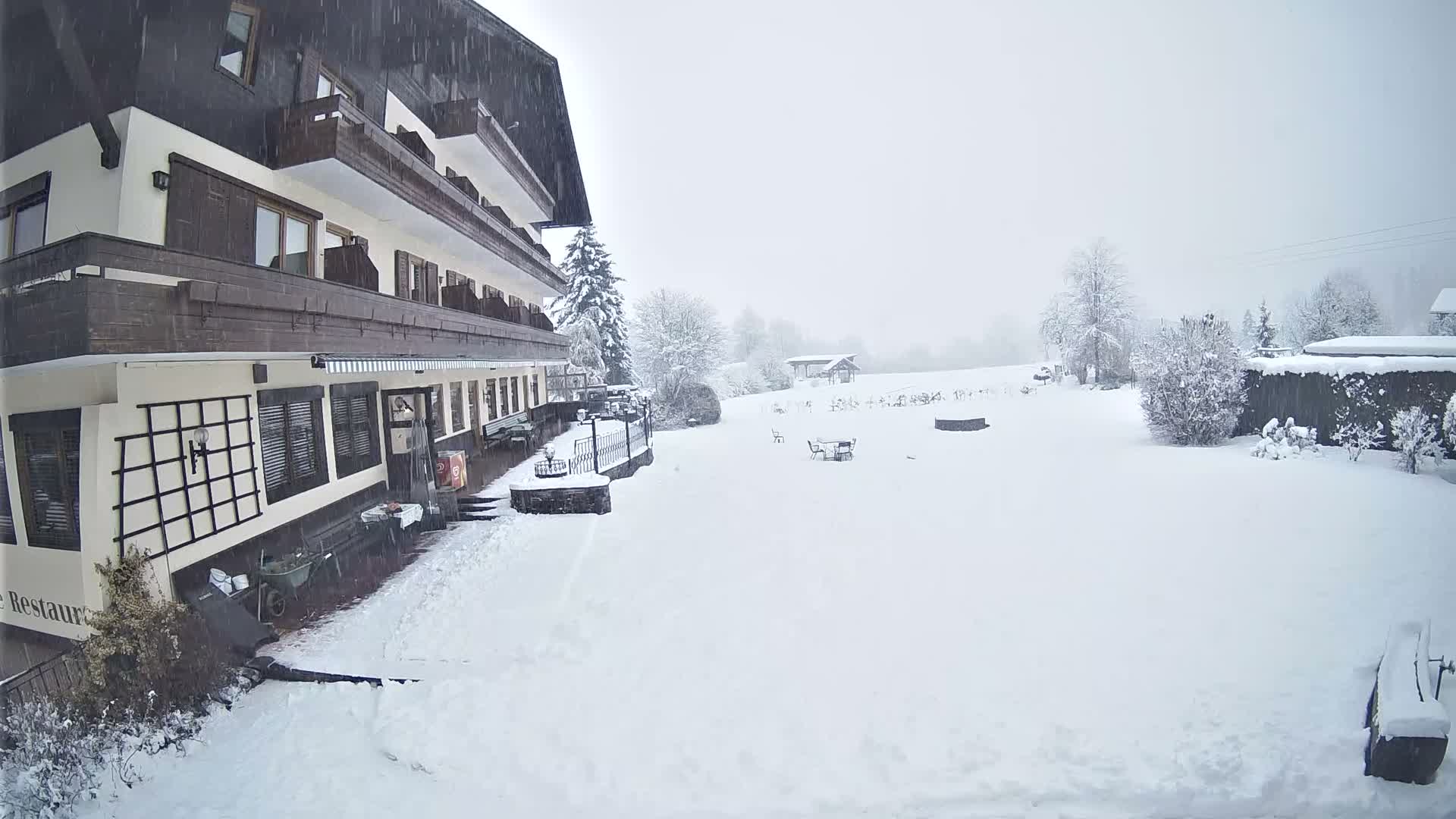 A large, multi-story building with balconies stands beside a vast, snow-covered landscape dotted with trees and a few distant structures, all under active heavy snowfall from a grey, overcast sky.