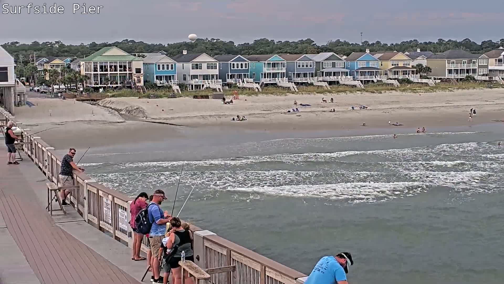Several people fish from a pier overlooking a beach with gentle waves and a row of colorful houses under an overcast sky.