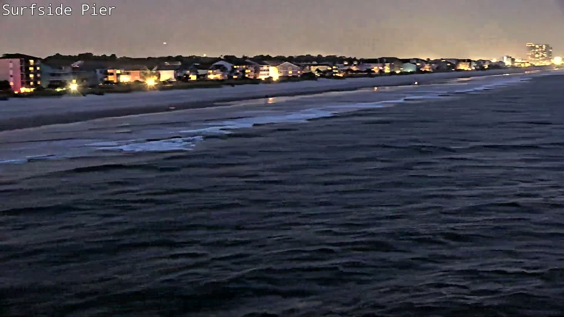 Several people fish from a pier overlooking a beach with gentle waves and a row of colorful houses under an overcast sky.