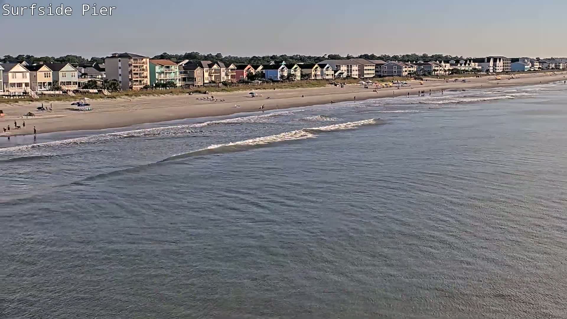 A nighttime view from a pier shows gently lapping waves, a sandy beach, and illuminated houses along the shore under a dark sky.