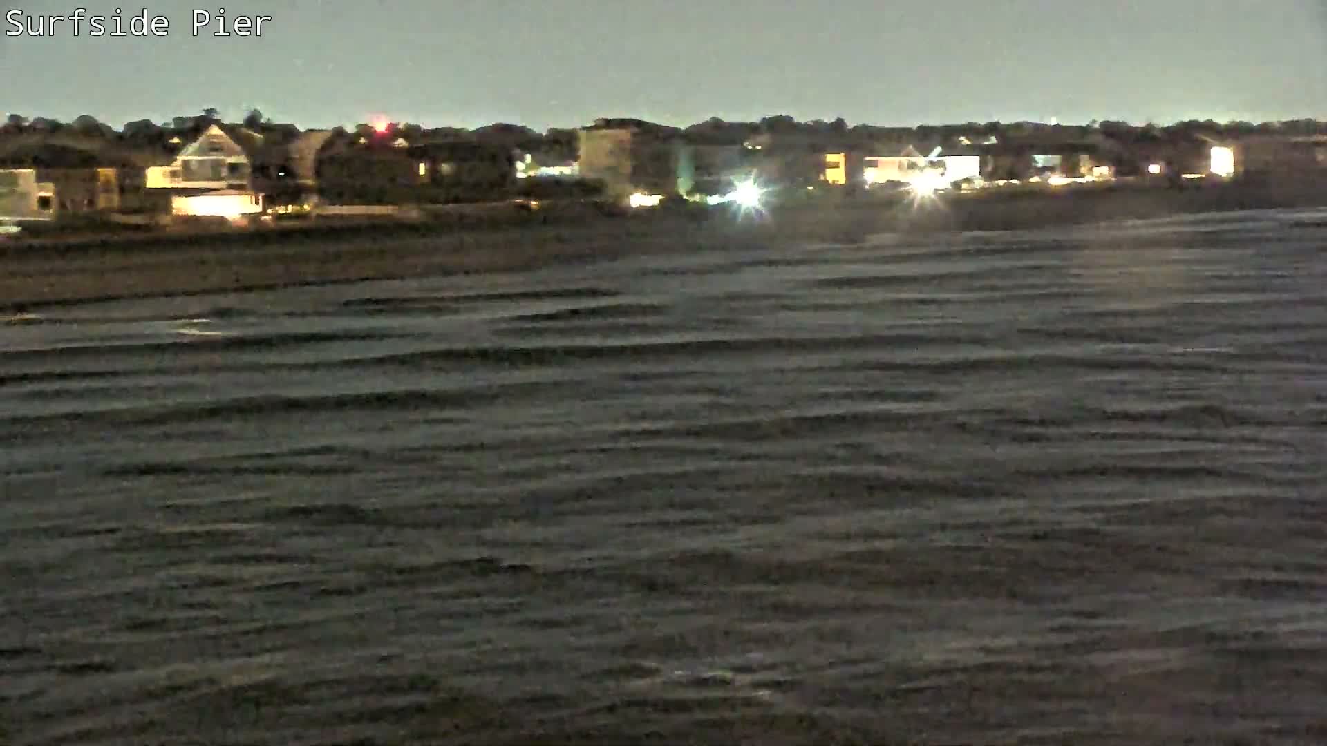 A choppy, dark body of water fills the foreground under an overcast night sky, with a distant illuminated coastline dotted with houses and buildings.