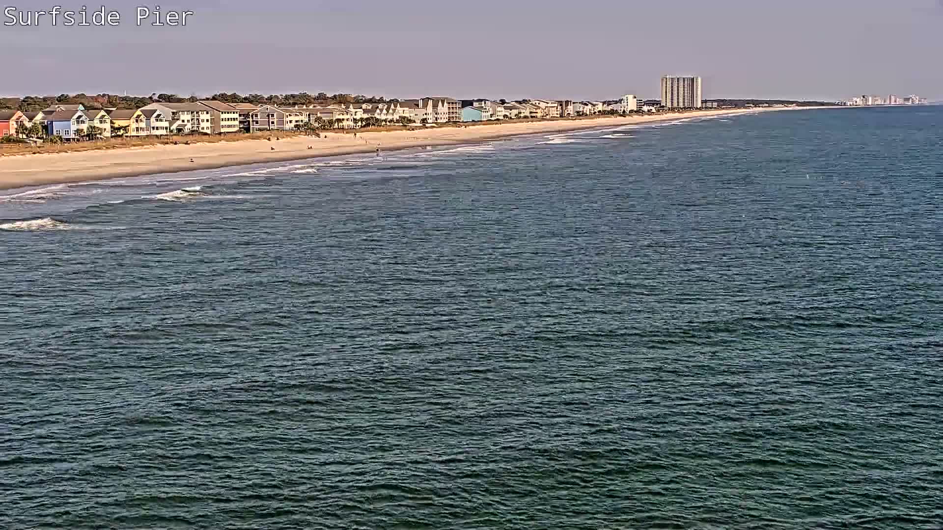 A choppy, dark body of water fills the foreground under an overcast night sky, with a distant illuminated coastline dotted with houses and buildings.