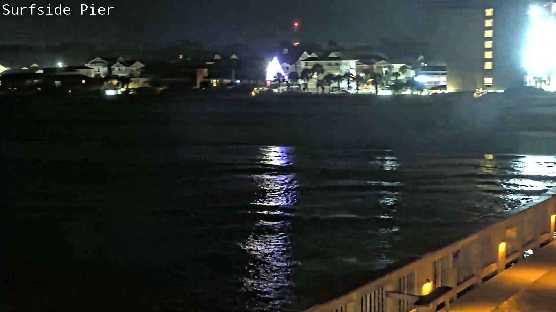 A nighttime scene from a pier shows dark, reflective water and distant illuminated buildings, including a prominent red flashing light, under rainy conditions.