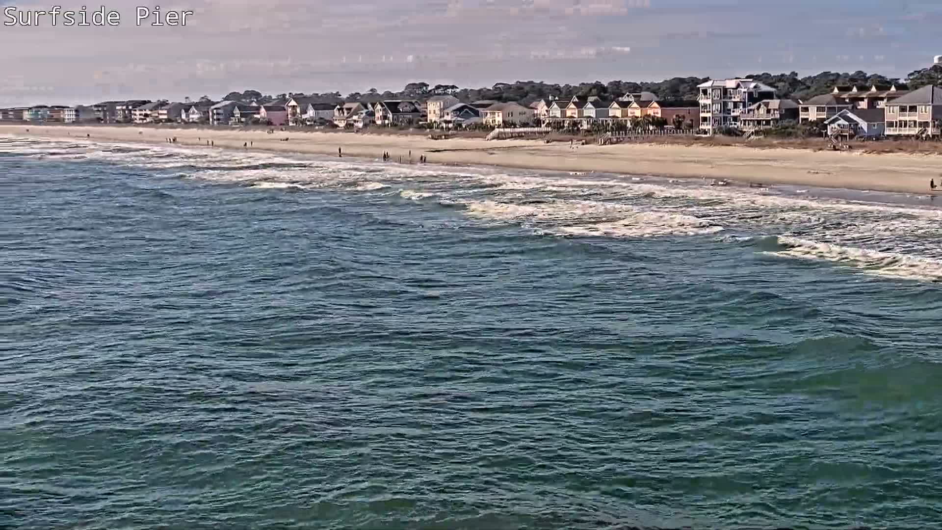 A nighttime scene from a pier shows dark, reflective water and distant illuminated buildings, including a prominent red flashing light, under rainy conditions.