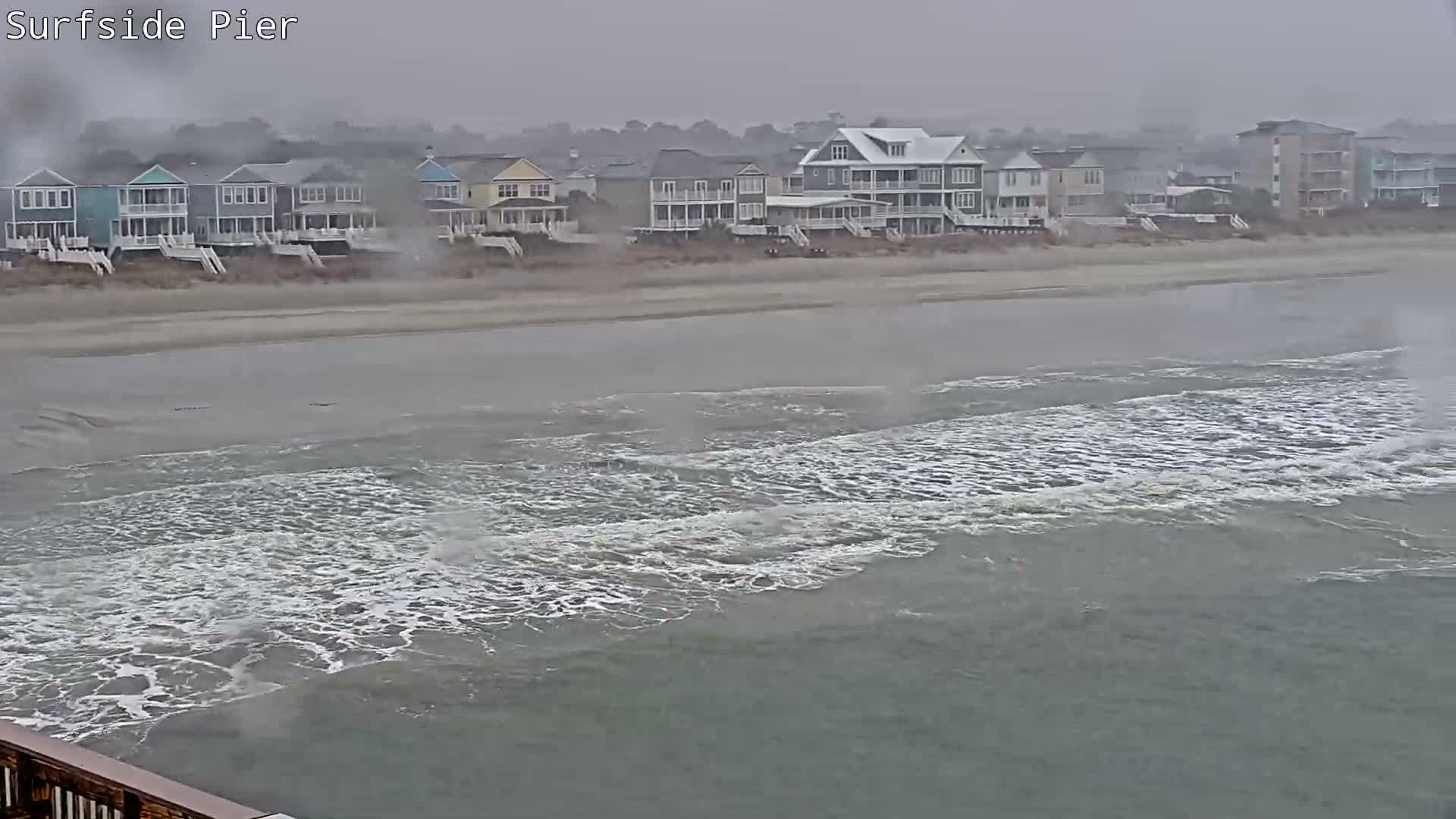 The image shows a stormy coastal scene with choppy waves breaking on a sandy beach lined with numerous multi-story houses under a heavily overcast and hazy sky.