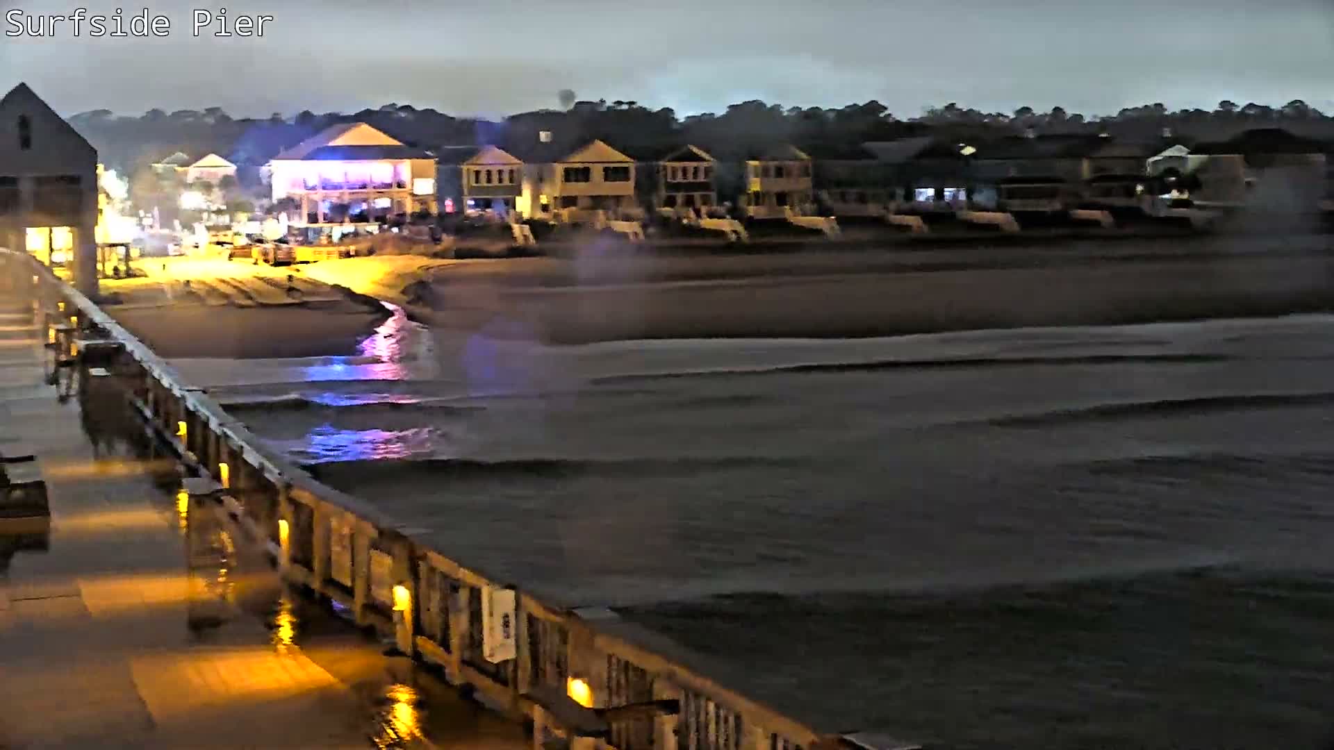 An evening scene from a damp, brightly lit pier overlooks a wavy ocean and a shoreline lined with illuminated buildings, all under a dark, overcast sky.