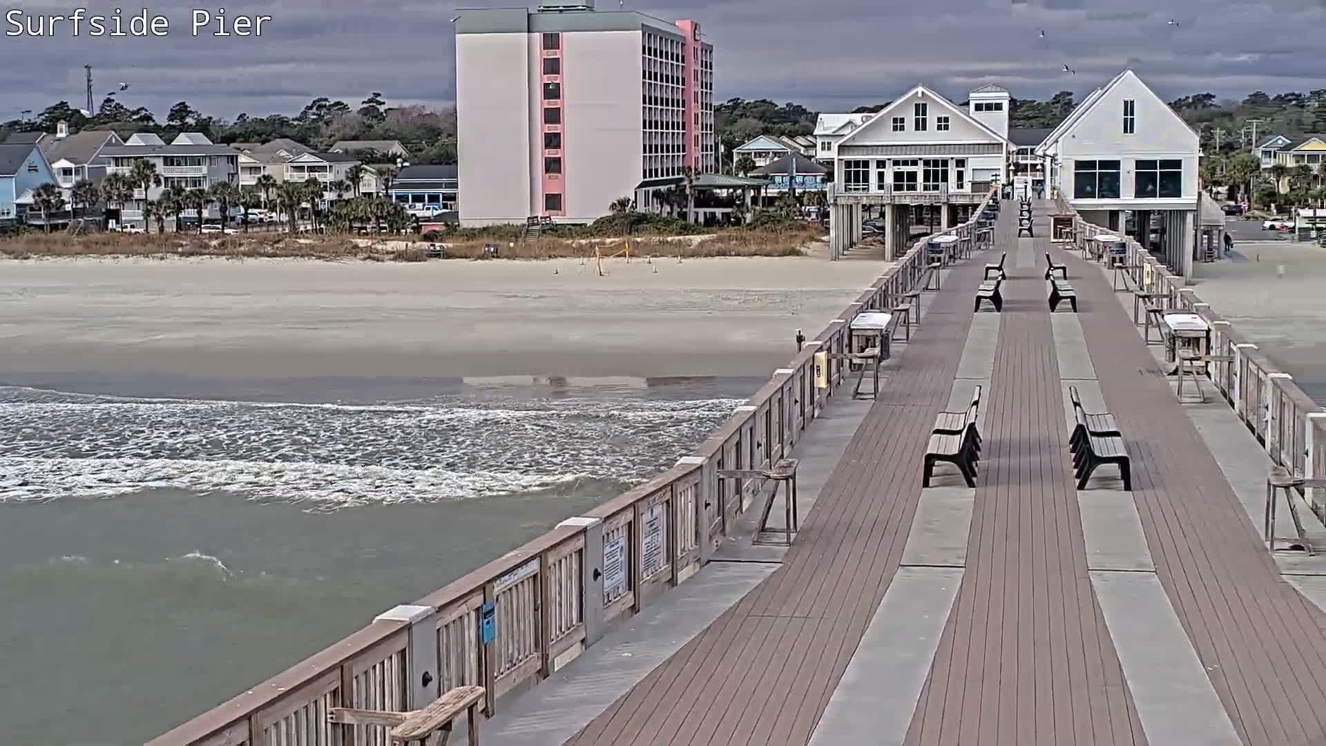 An evening scene from a damp, brightly lit pier overlooks a wavy ocean and a shoreline lined with illuminated buildings, all under a dark, overcast sky.