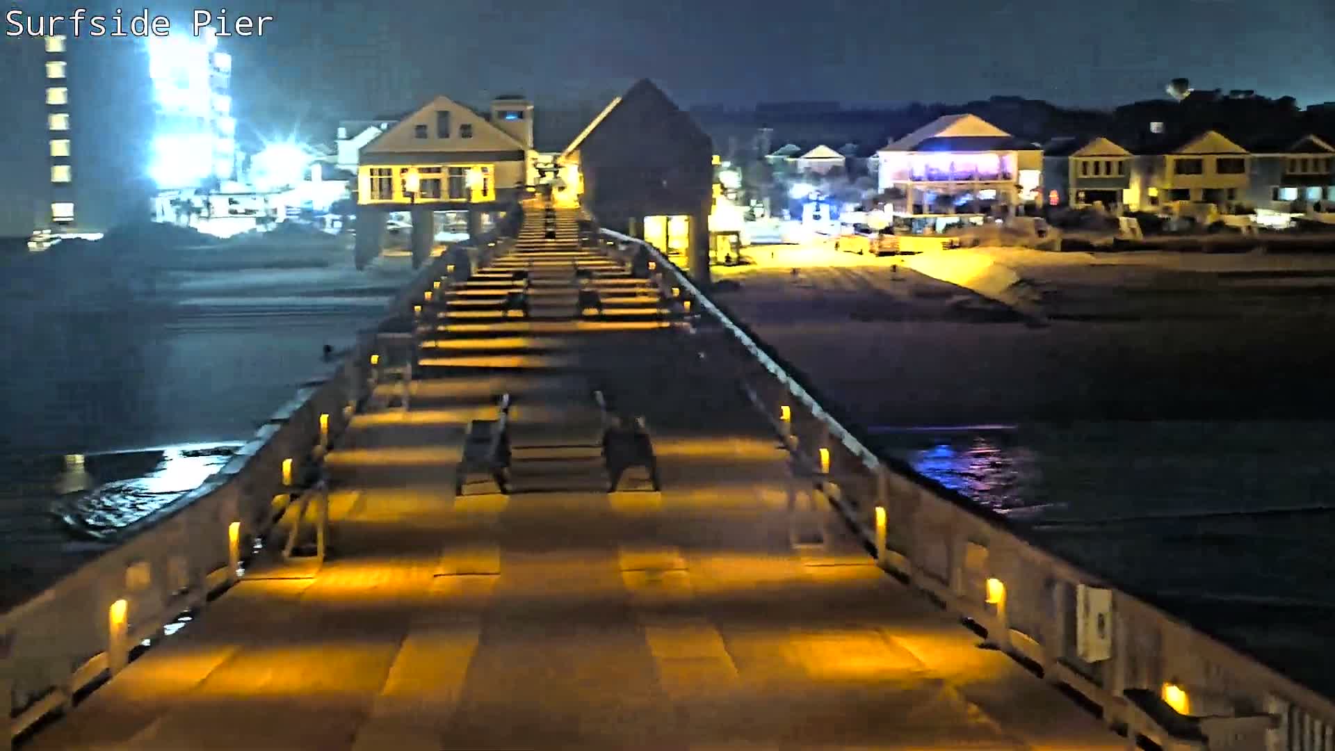An evening scene from a damp, brightly lit pier overlooks a wavy ocean and a shoreline lined with illuminated buildings, all under a dark, overcast sky.