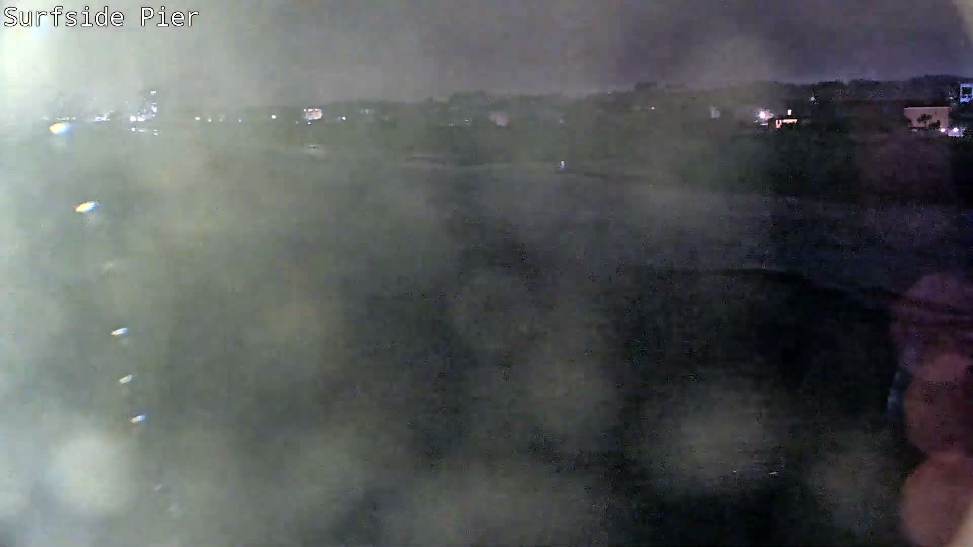 A heavily obscured and blurry night view of a coastal area, with distant lights appearing diffused through dense fog or rain on the camera lens.