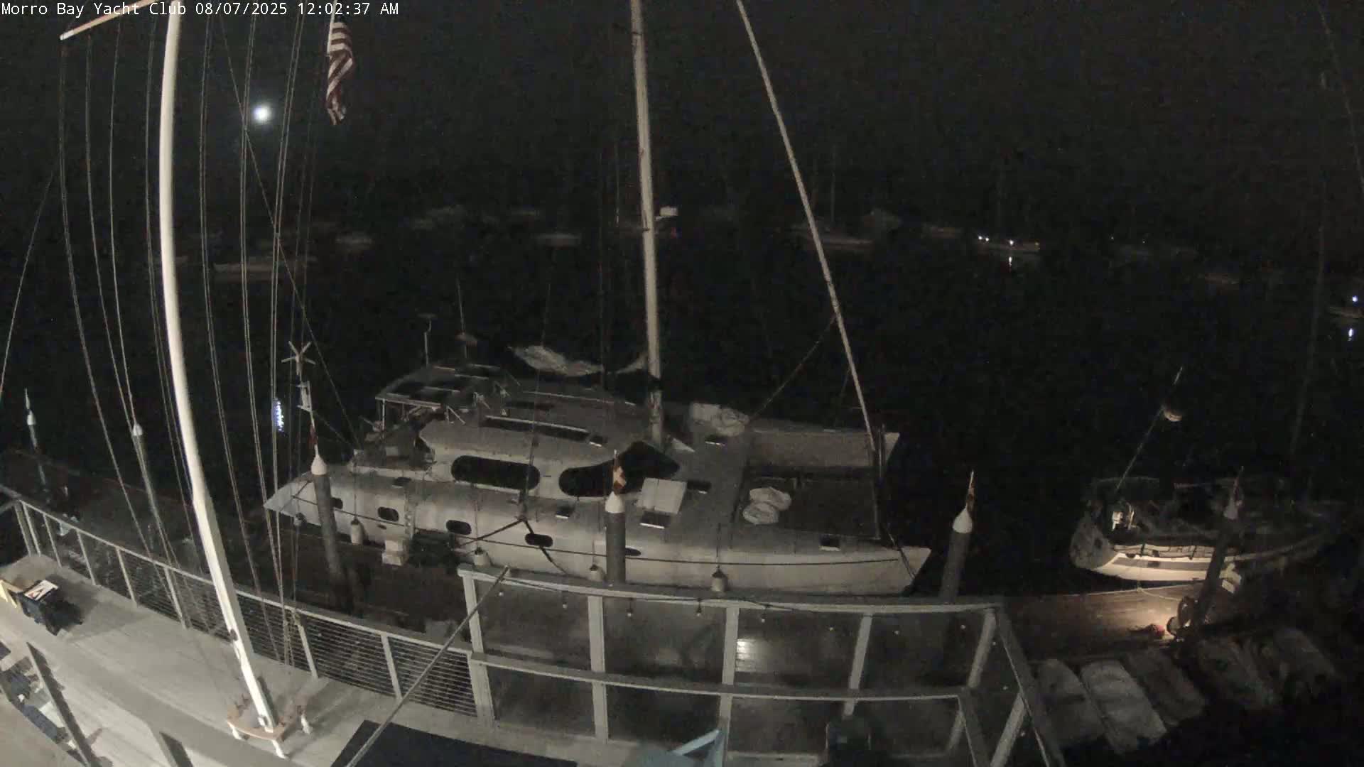 A nighttime, high-angle view shows several boats docked at a marina under a dark sky with a visible moon.