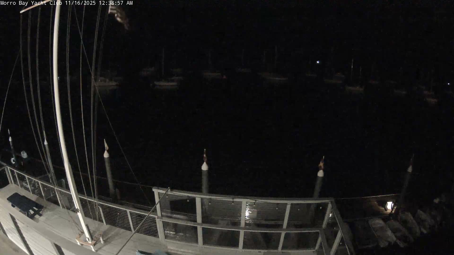 A clear night view of a marina captures the mast and rigging of a foreground boat, with numerous other vessels dimly visible on the calm, dark water.