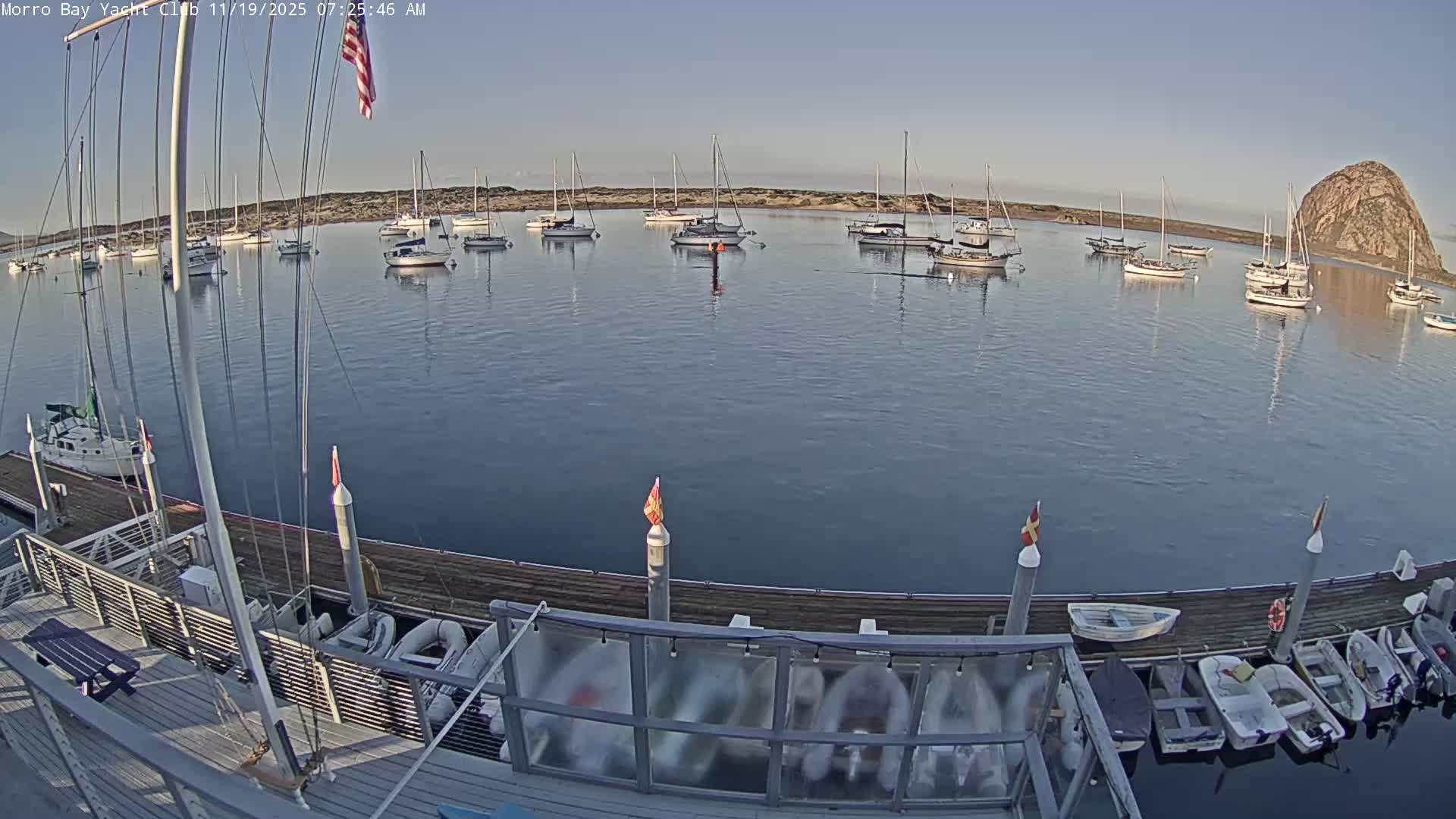A clear night view of a marina captures the mast and rigging of a foreground boat, with numerous other vessels dimly visible on the calm, dark water.