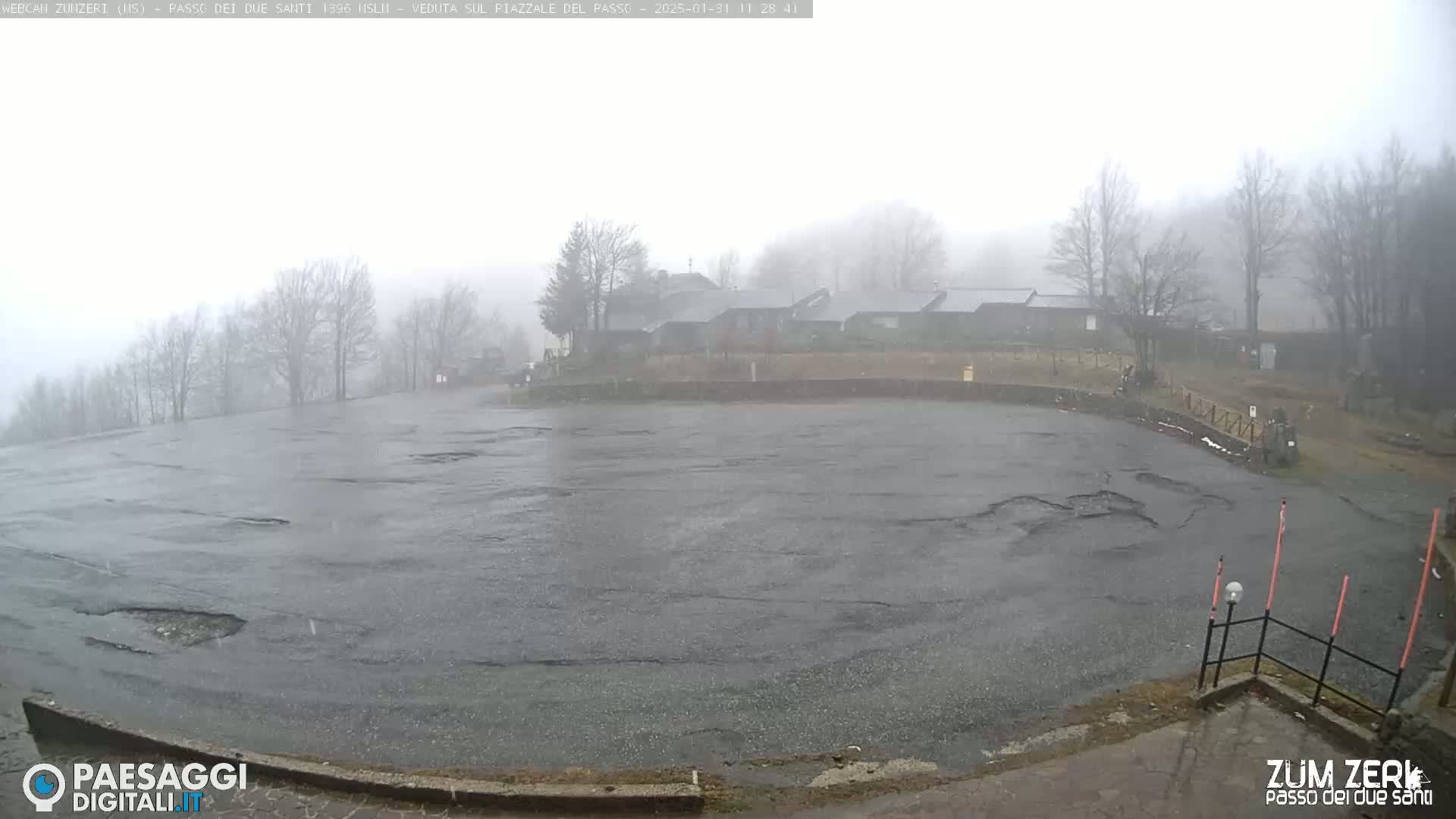 A large, wet, empty parking lot is seen under a heavy fog with a few buildings visible in the distance.